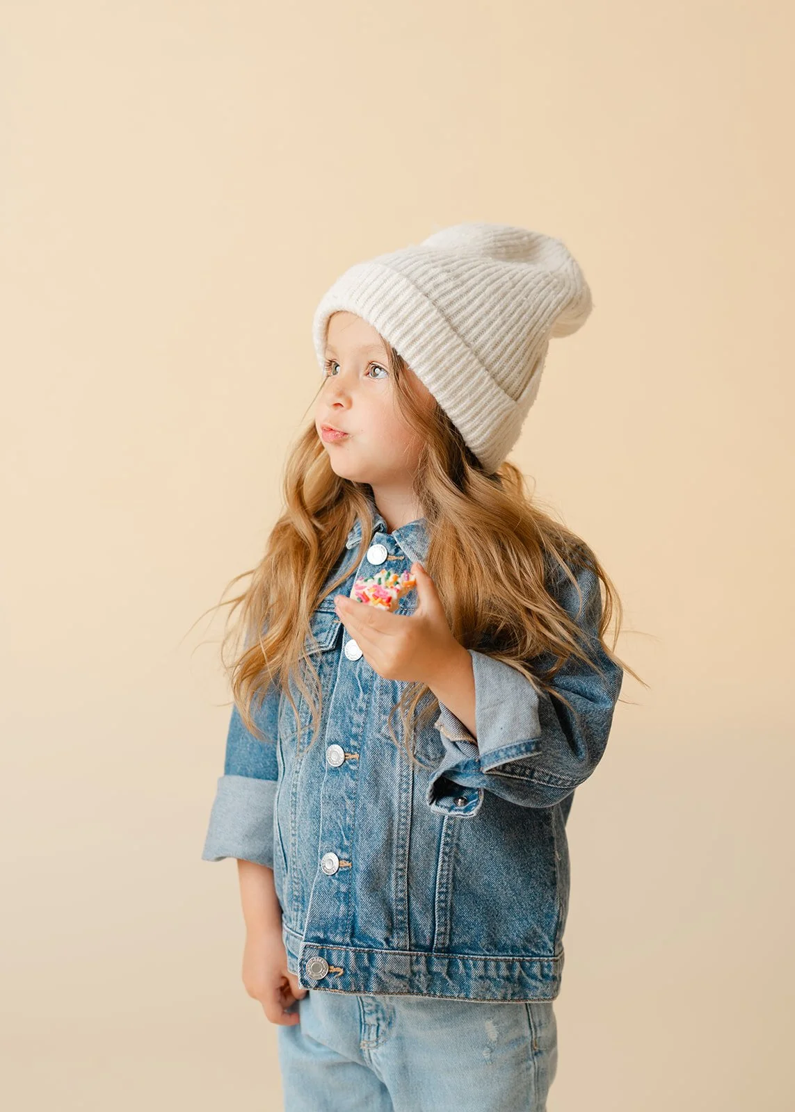 A young girl with long red hair wearing a white knit beanie and denim jacket holding a colorful sprinkled donut.