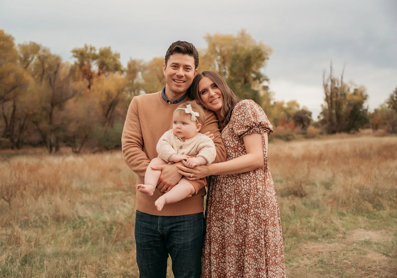 A family of three outdoors in autumn, with trees in fall foliage in the background. The man is holding a baby girl, and the woman is standing next to them smiling.