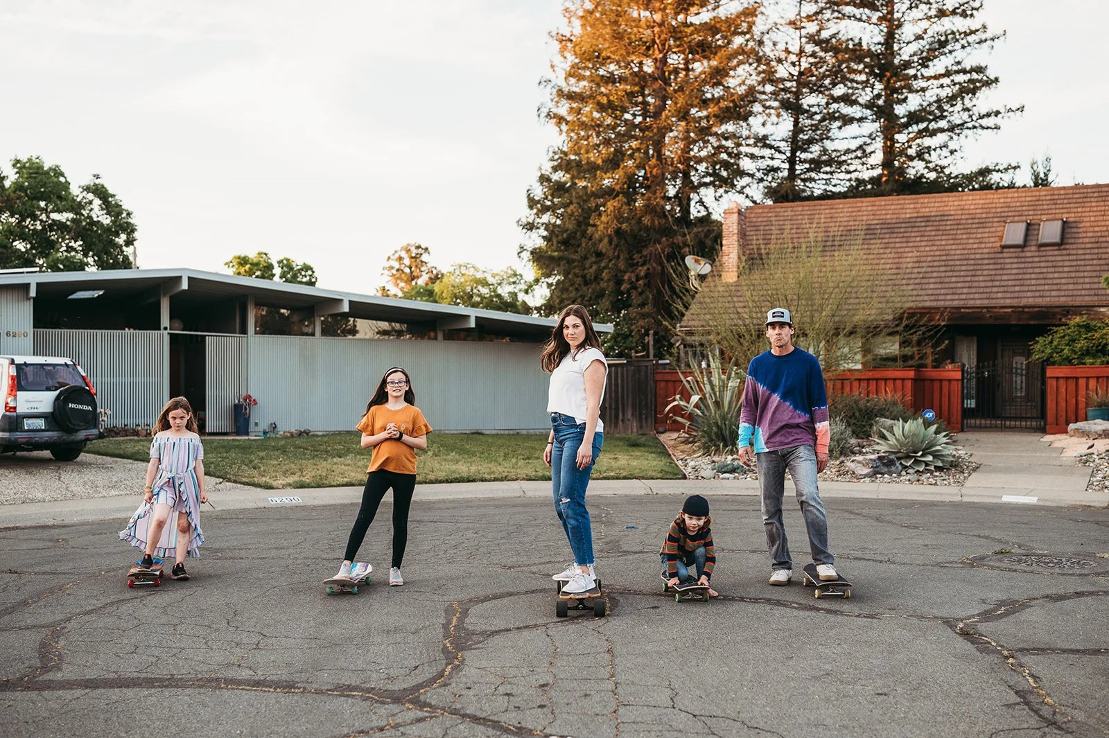Family of six skateboarding in a neighborhood street during sunset. Adult woman and man along with four children, some on skateboards, in front of houses with trees and a clear sky background.