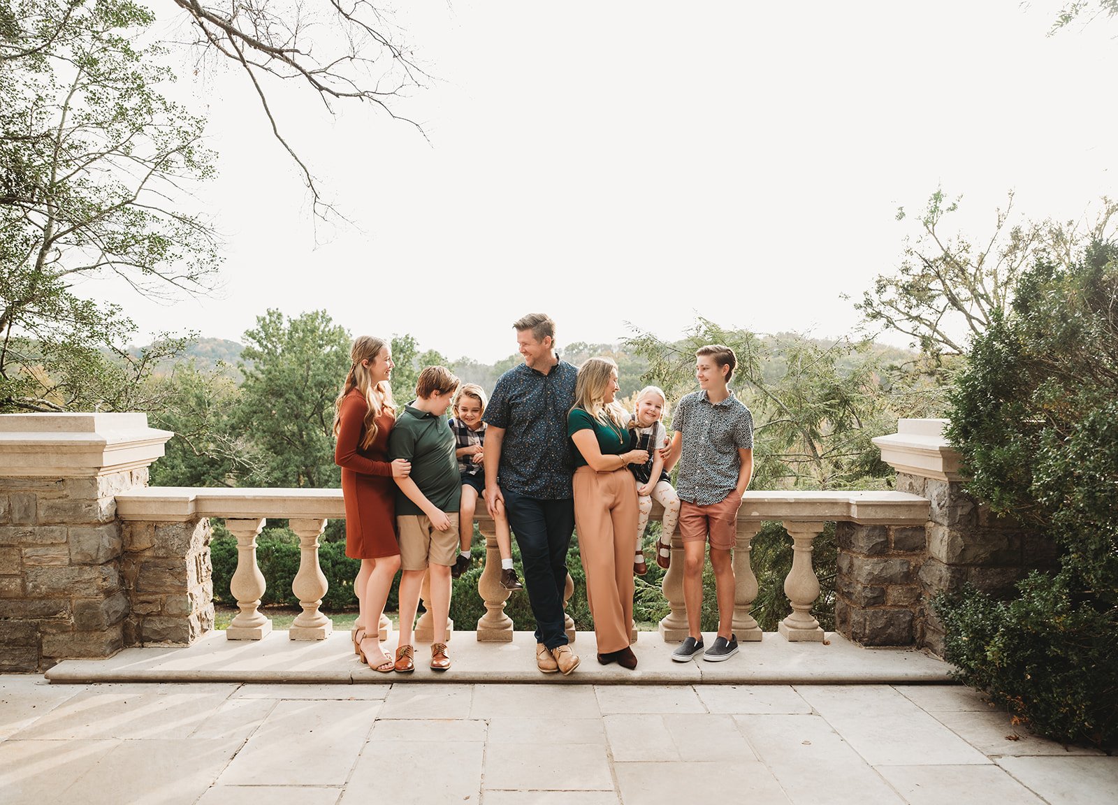A family of eight standing on a stone balcony with balustrades, surrounded by trees and greenery, smiling and enjoying each other's company.