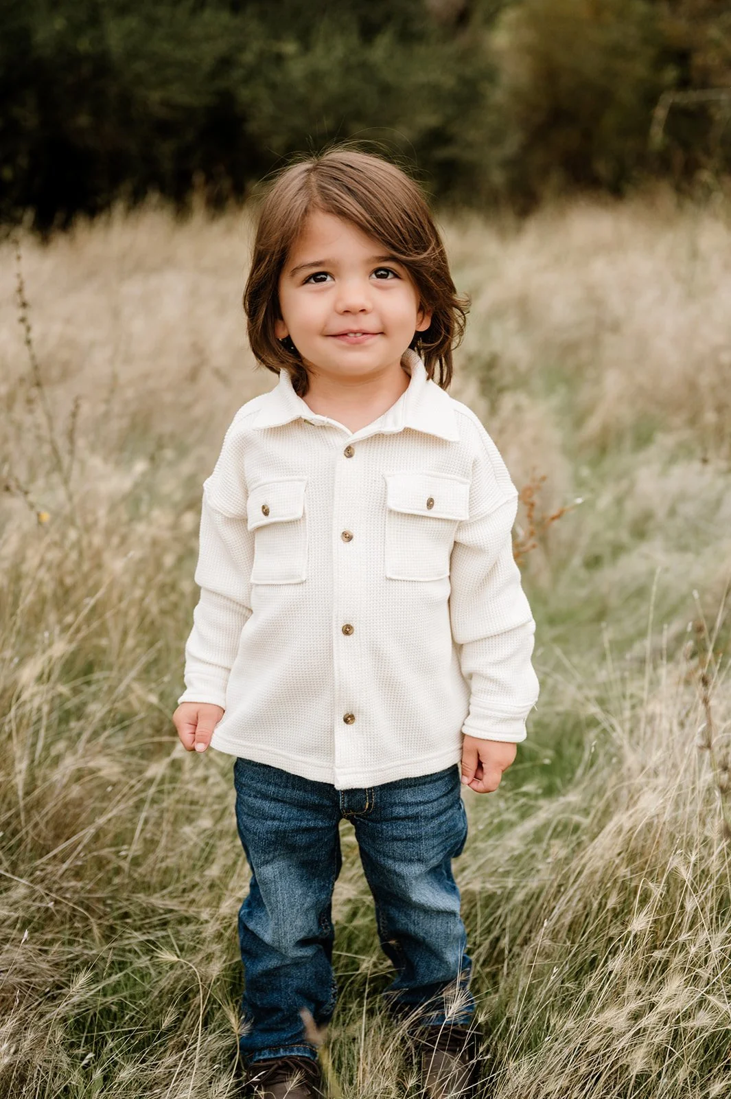 A young child with brown hair, wearing a white button-up shirt and jeans, standing in a grassy field with trees in the background.