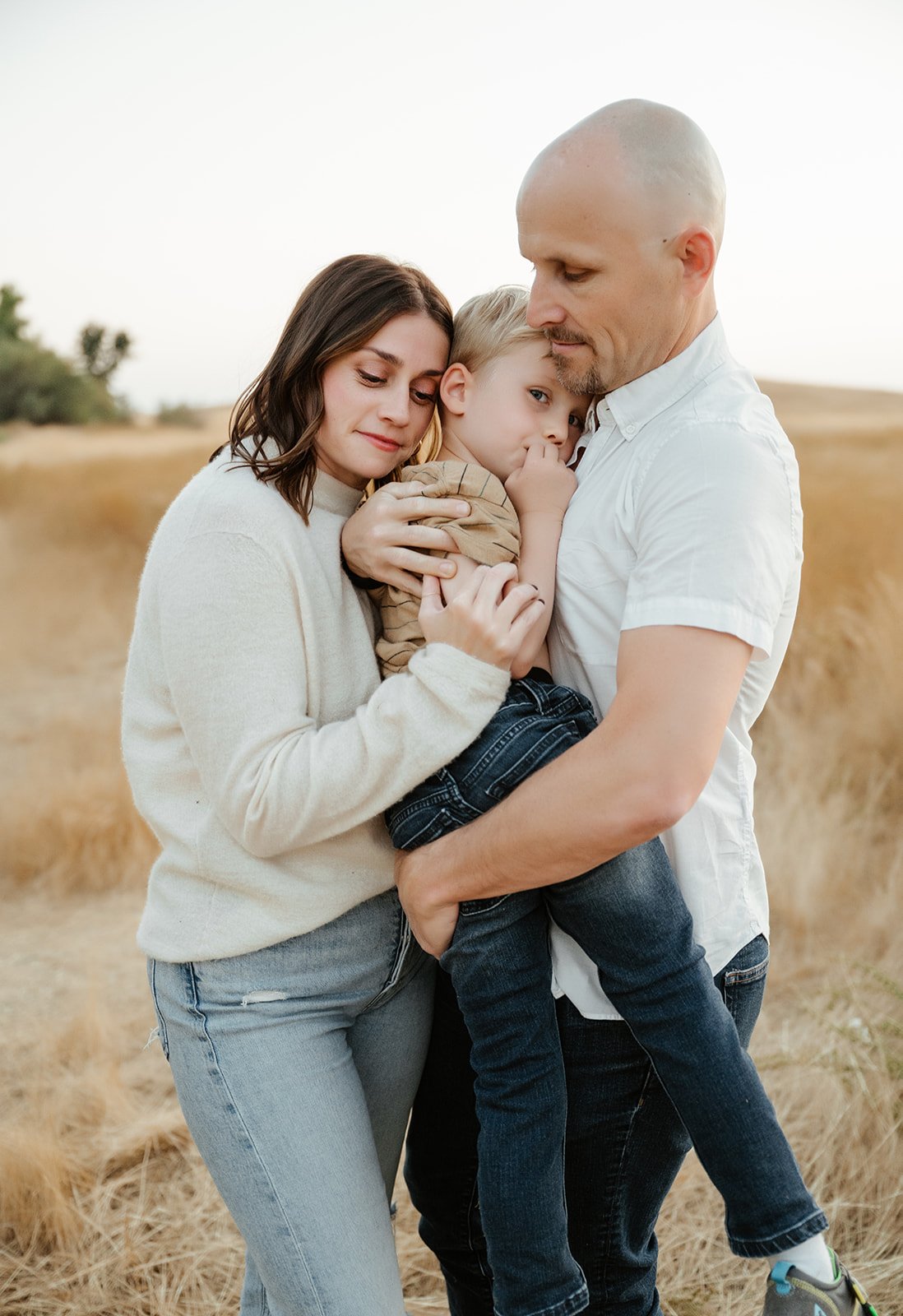 A family of three embracing outdoors in a field with dry grass, showing a woman and man holding a young boy.