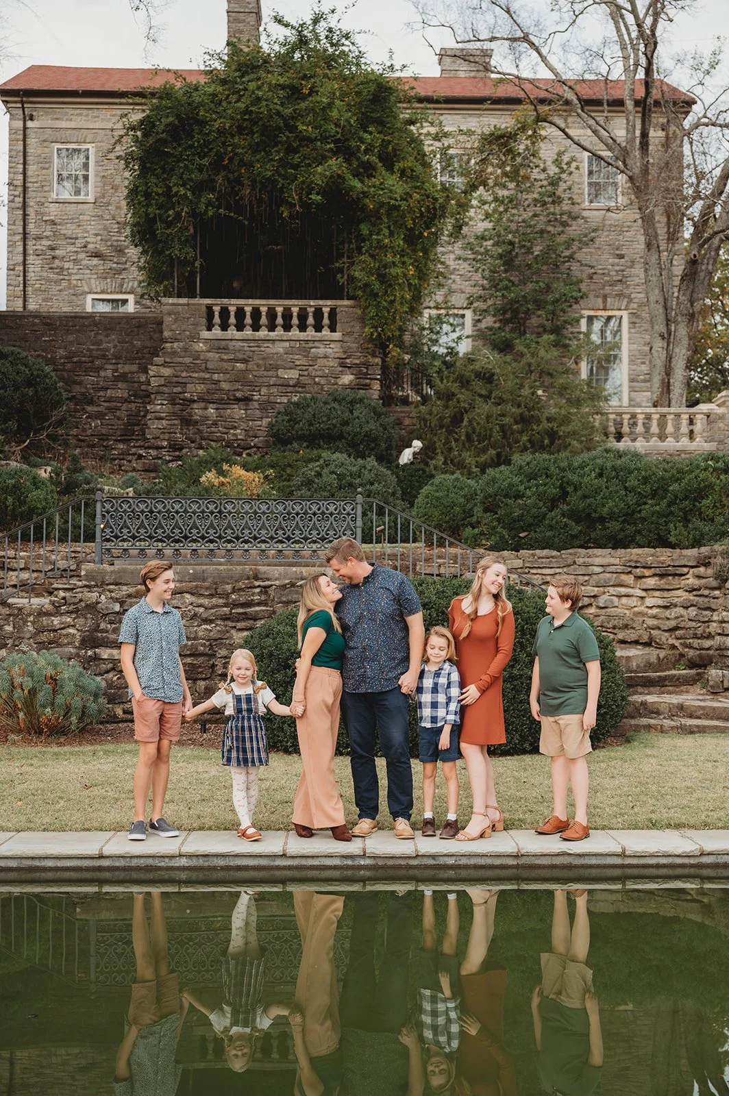 A family of seven stands by a pool in a backyard with a stone house and garden. The adults and children are smiling and holding hands, with some looking at each other and some looking at the camera.