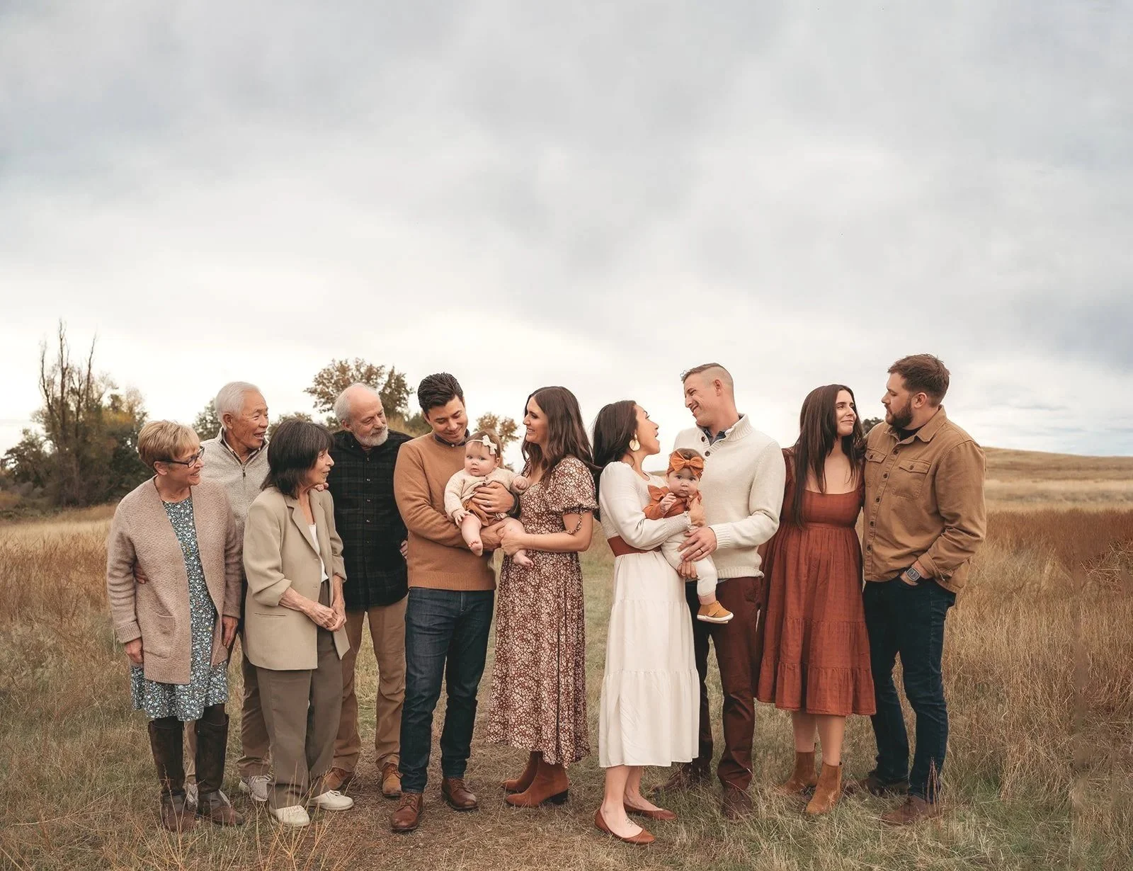 Family gathering outdoors on a cloudy day, standing in a line across a grassy field, including elderly, adults, and children.