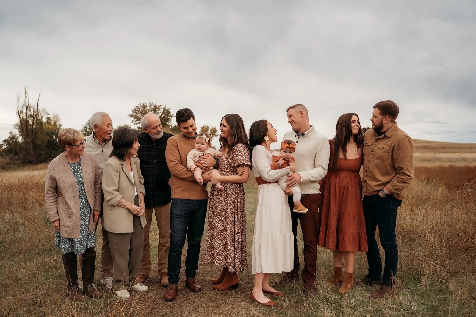 A multi-generational family standing together outdoors in a field with cloudy sky, including grandparents, parents, children, and babies, dressed in casual fall clothing.