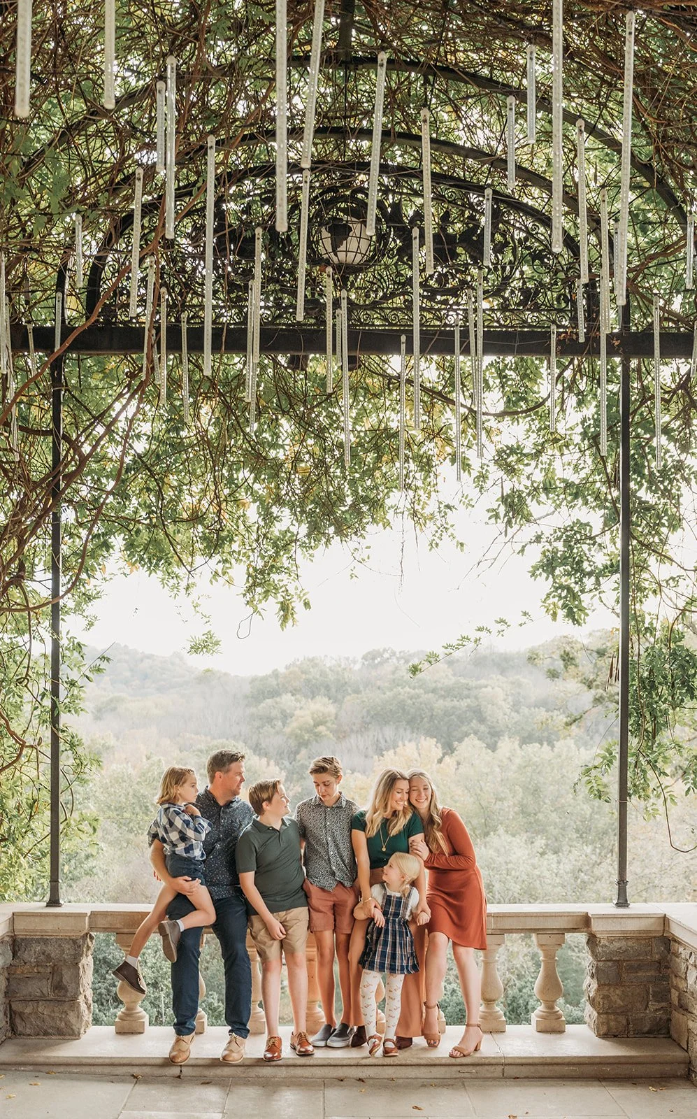 Family of seven standing on a stone balcony under a pergola covered in plants, with a scenic view of trees and hills in the background.