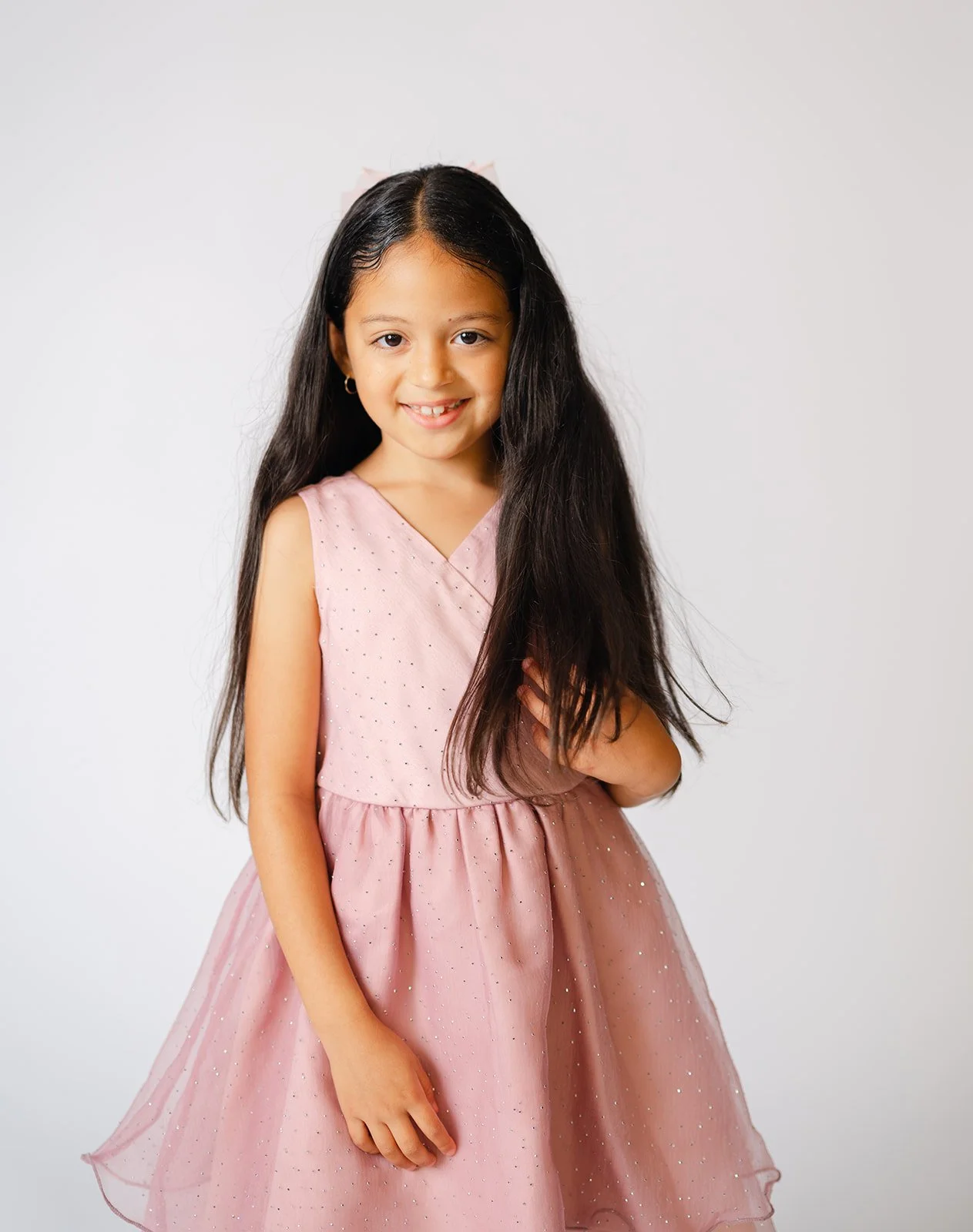 Young girl in a pink dress with long dark hair smiling at the camera, against a plain white background.