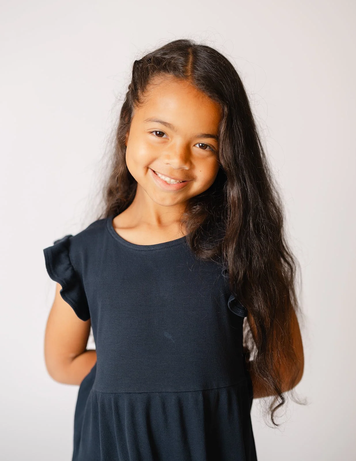 A young girl with long dark hair smiling and wearing a black dress against a plain background.