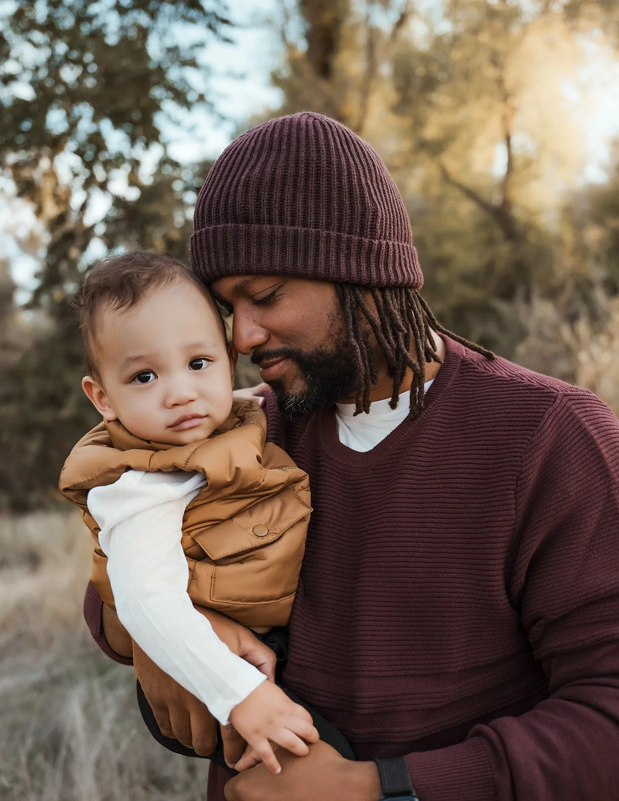 A man with dreadlocks wearing a maroon beanie and maroon sweater holds a young child with light skin, brown hair, and big eyes, in an outdoor setting. The child looks at the camera with a serious expression. The background features trees with sunligh