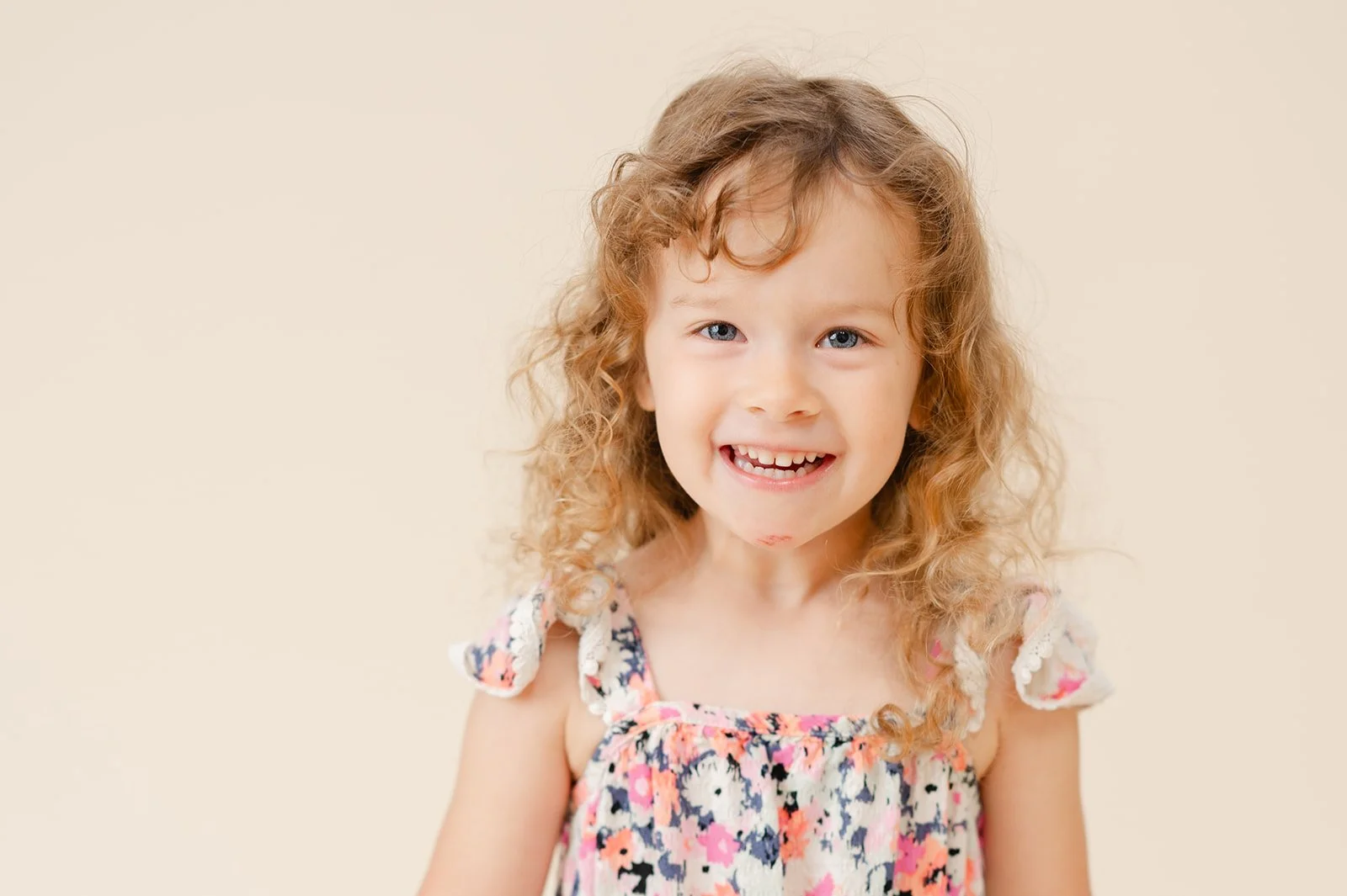 Smiling young girl with curly hair wearing a floral dress against a plain background.