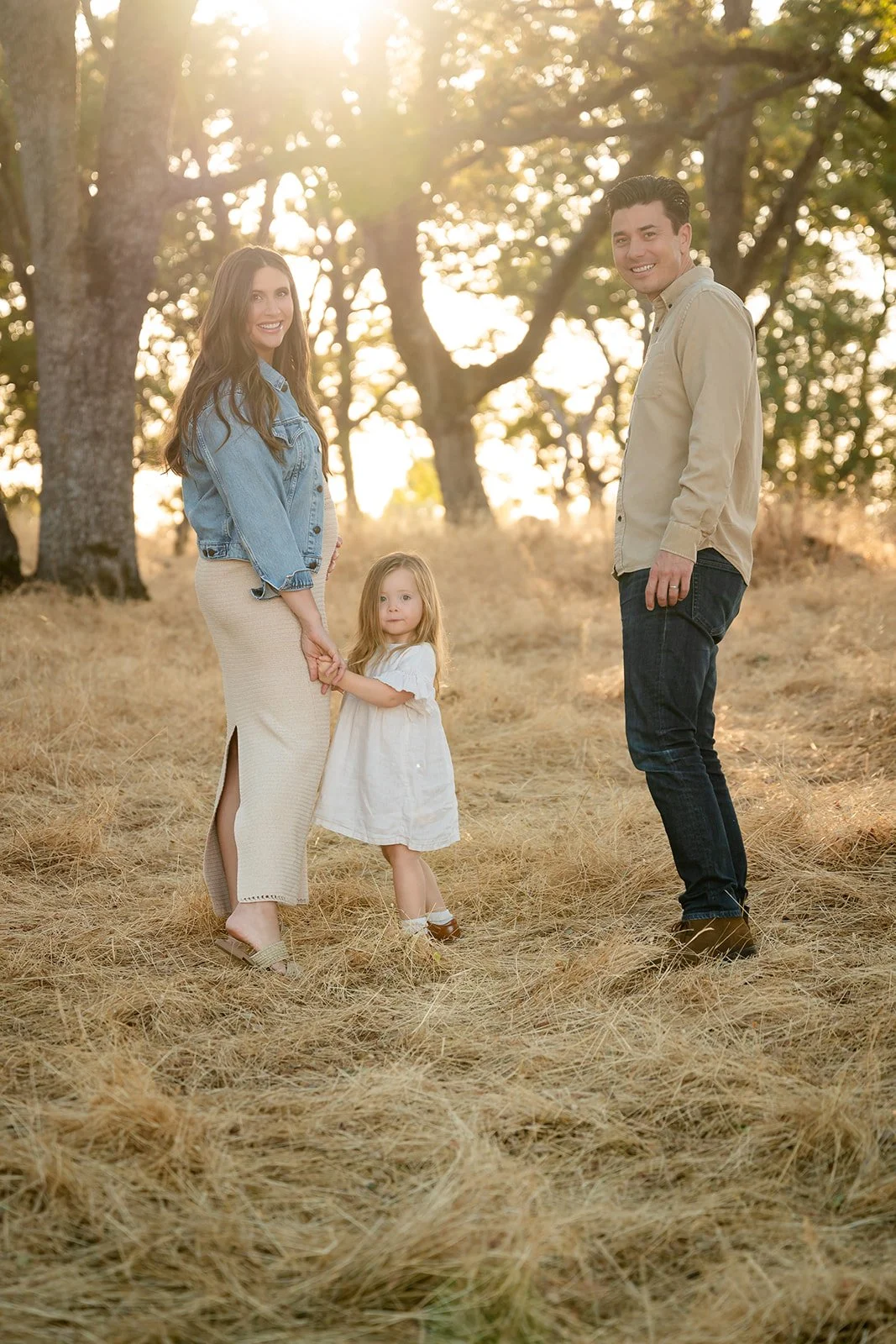 A family of three, including a woman, a man, and a young girl, standing outdoors in a field with tall grass and trees in the background during sunset or late afternoon.