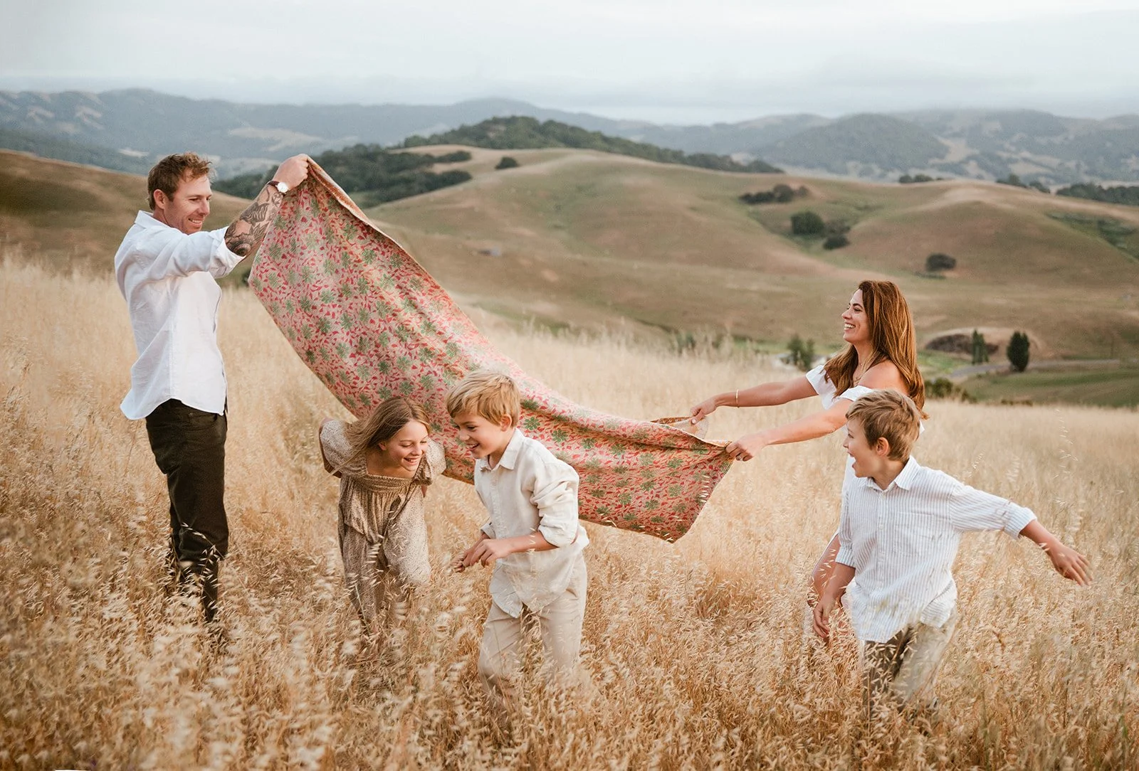 A family of five enjoying time in a field of tall, golden grass with rolling hills in the background. The father and mother are holding a colorful blanket as the children play and laugh beneath it.