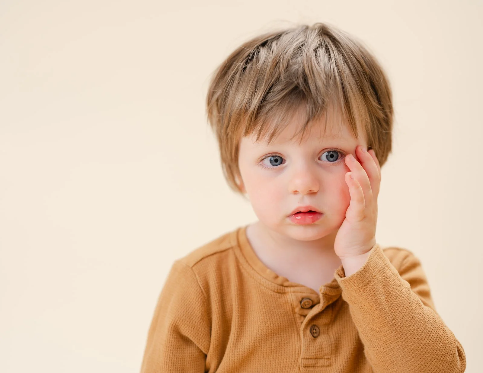 A young Caucasian boy with light brown hair and blue eyes, wearing a tan long-sleeve shirt, touching his face with his hand, standing against a plain beige background.