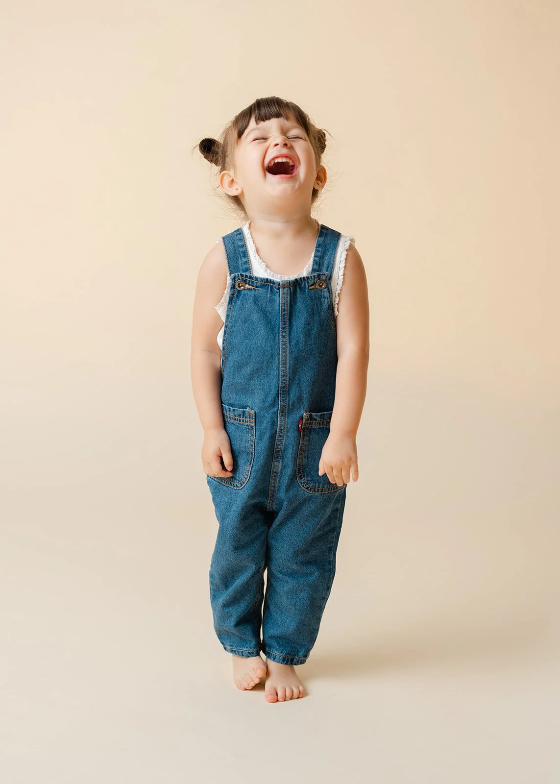 Young girl with pigtails laughing, wearing denim overalls and standing barefoot against a beige background.