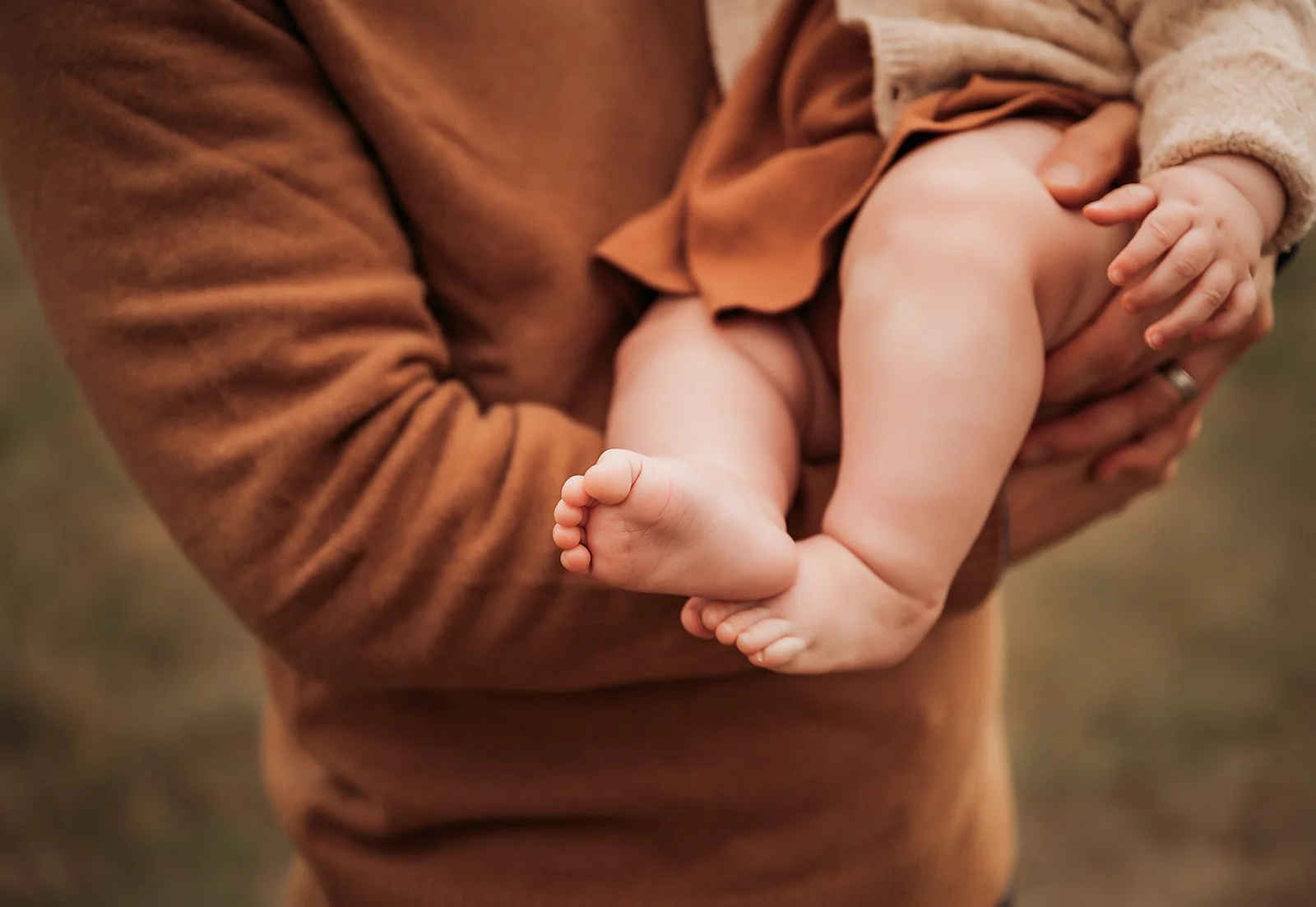 Close-up of a person holding a baby with focus on the baby's legs and feet, in an outdoor setting.