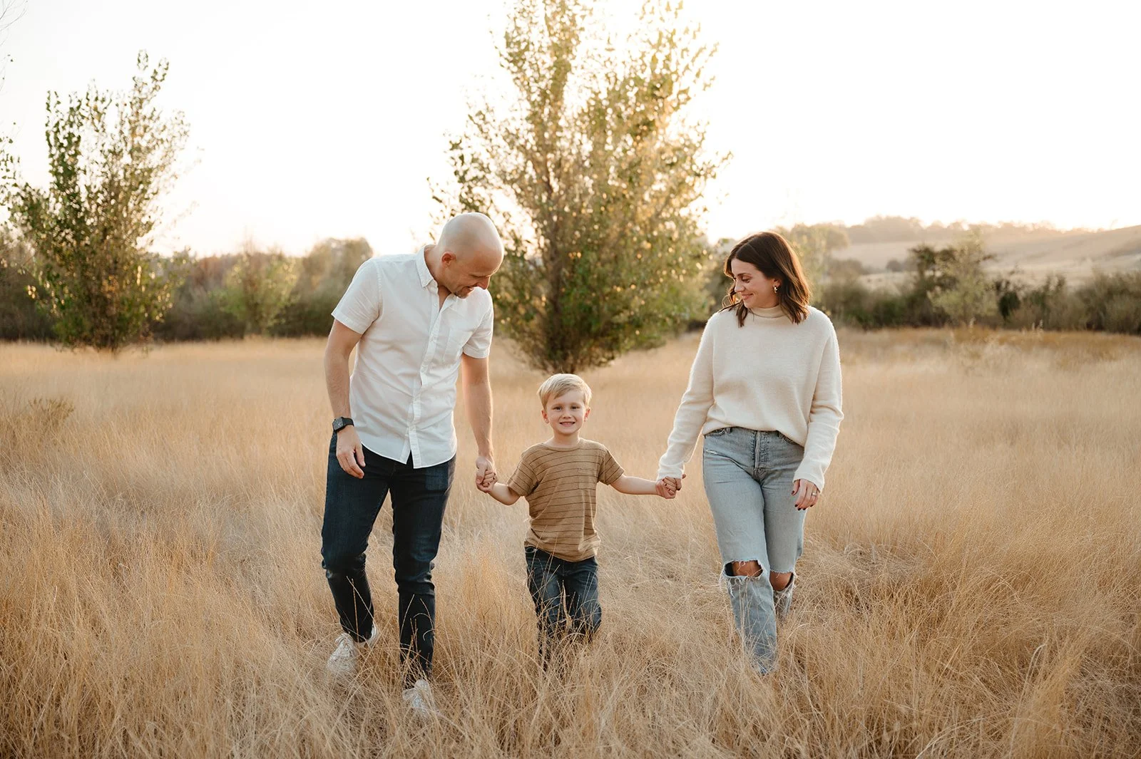 A family of three walking hand in hand through a grassy field during sunset, with trees in the background.