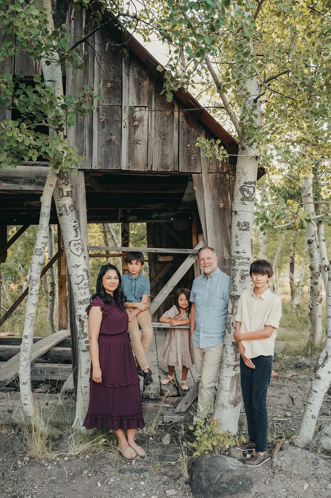 Family of five standing outdoors near a rustic wooden treehouse and trees, with two boys, a girl, a woman, and a man.