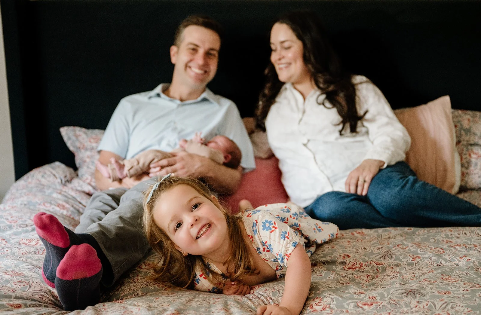 A family of four on a bed, including a man holding a newborn, a woman sitting beside him, a smiling young girl lying on her stomach in front, and a baby in his father's arms.