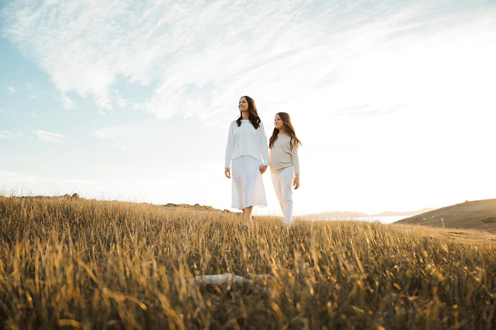 A woman and a girl walking through a grass field at sunset, holding hands, wearing white clothing.
