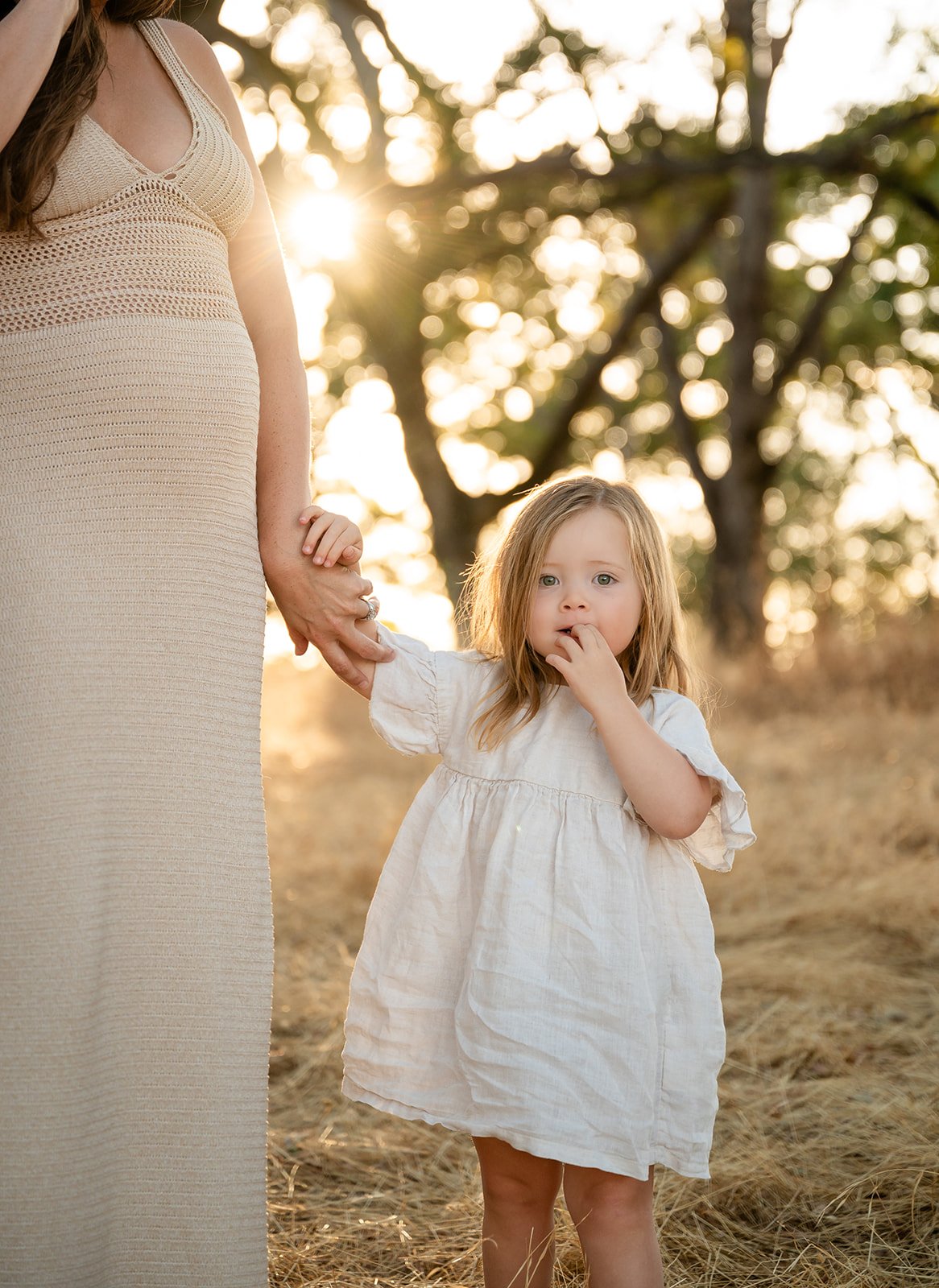 A young girl with red hair and green eyes standing outdoors in a field during sunset, wearing a white dress, holding her mother's hand, with a slightly worried or curious expression.