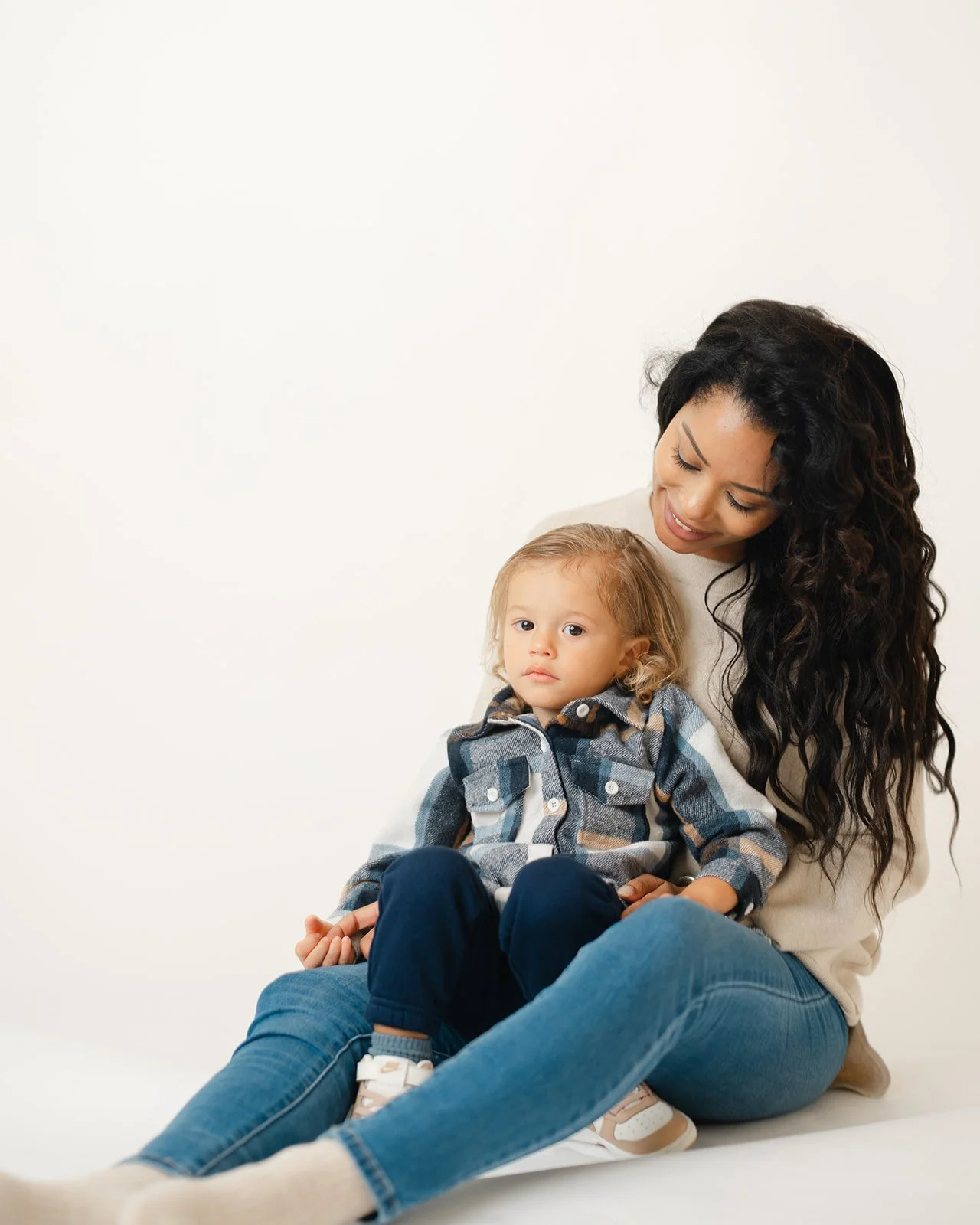 A woman with long curly dark hair sitting with a young child on her lap against a plain white background.