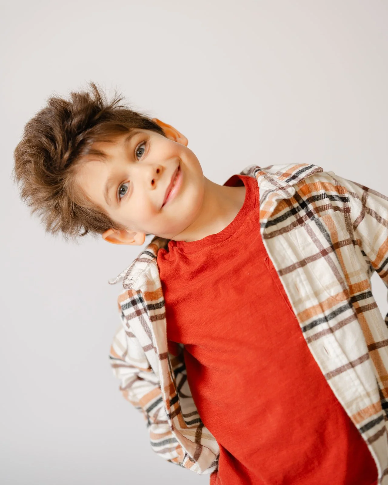 Young boy with light brown hair, blue eyes, smiling, wearing a red shirt and plaid jacket, standing against a plain white background.