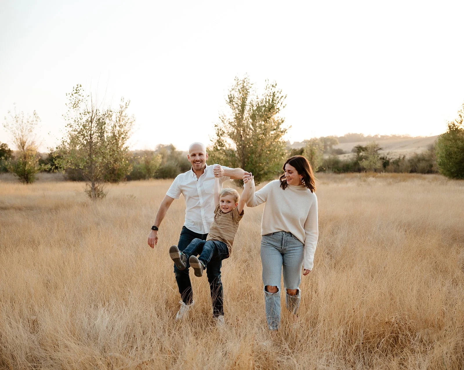A family of three, a father, mother, and young son, walking and playing in a field of tall, dry grass during sunset, holding hands and smiling.