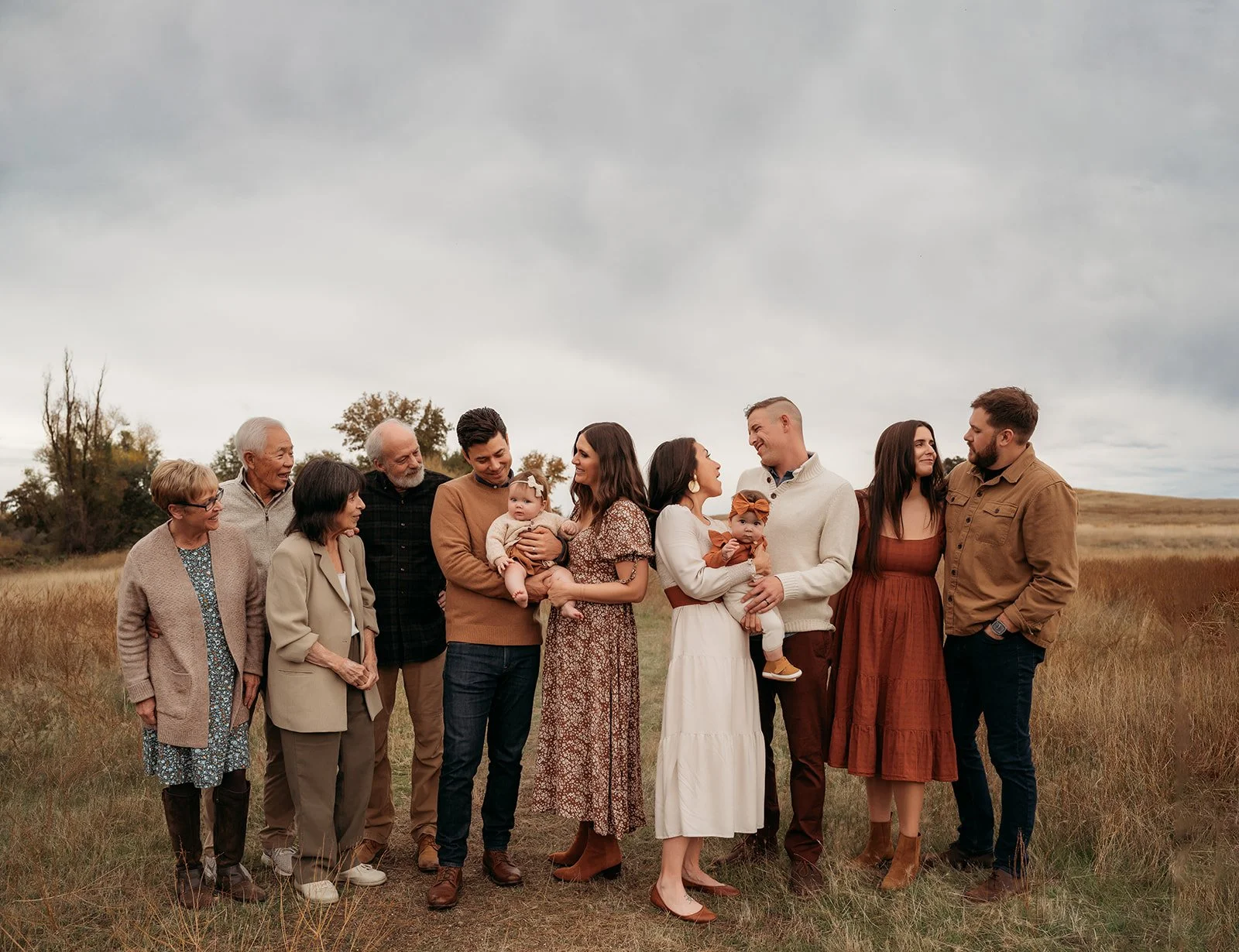 Family group standing outdoors in a grassy field under a cloudy sky, dressed in casual Autumn attire, smiling and interacting.