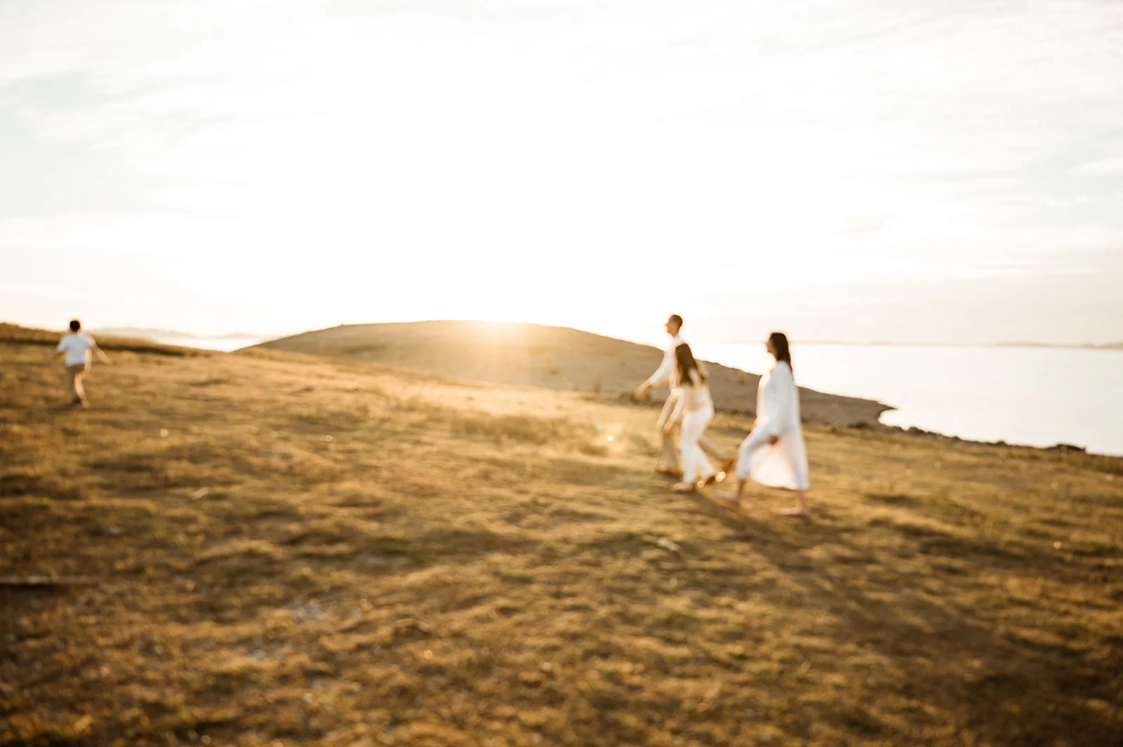 Four people walking on a grassy hill near a body of water during sunset, with the sun low in the sky illuminating the scene.