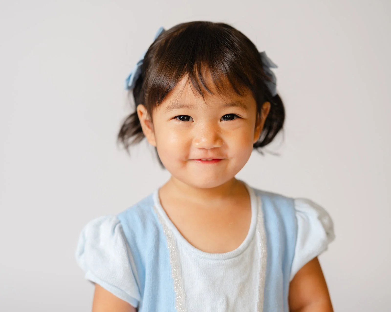 Young girl with short black hair and bows, smiling, wearing a light blue dress with white accents, against a plain light background.