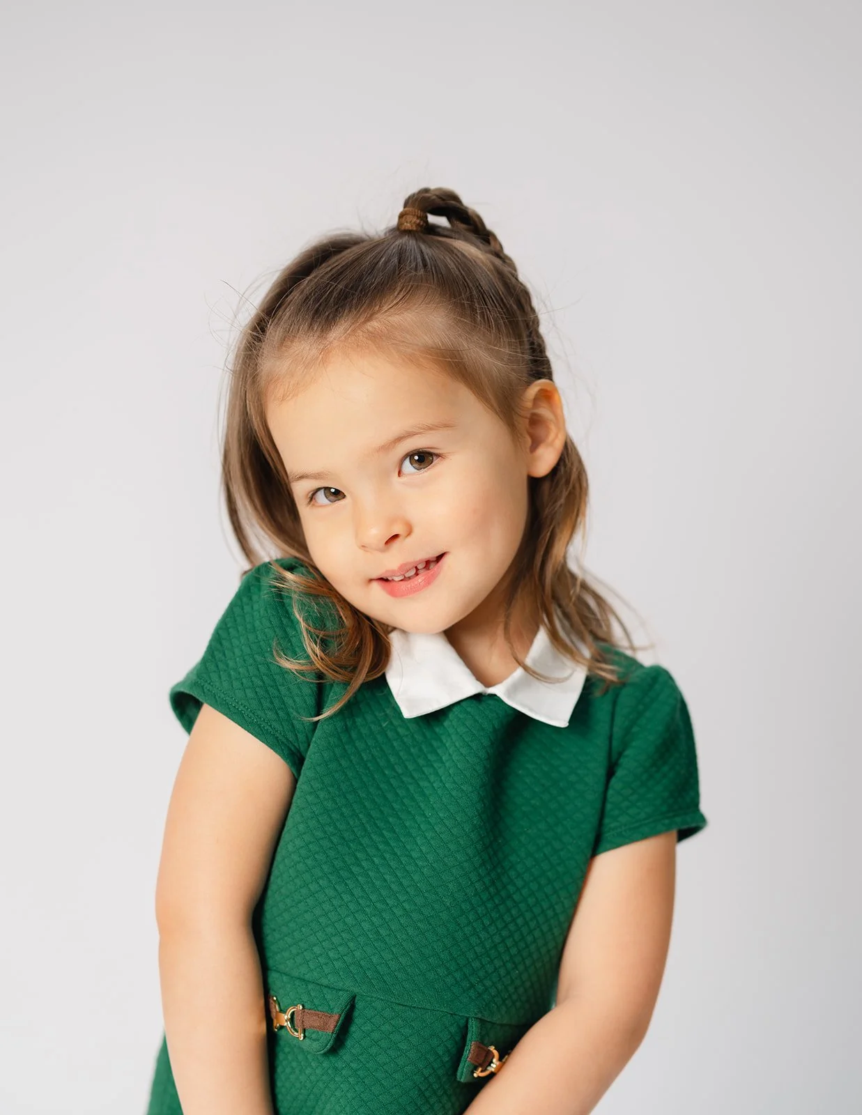 A young girl with light brown hair styled with a half braid, wearing a green dress with a white collar, standing against a plain light gray background.