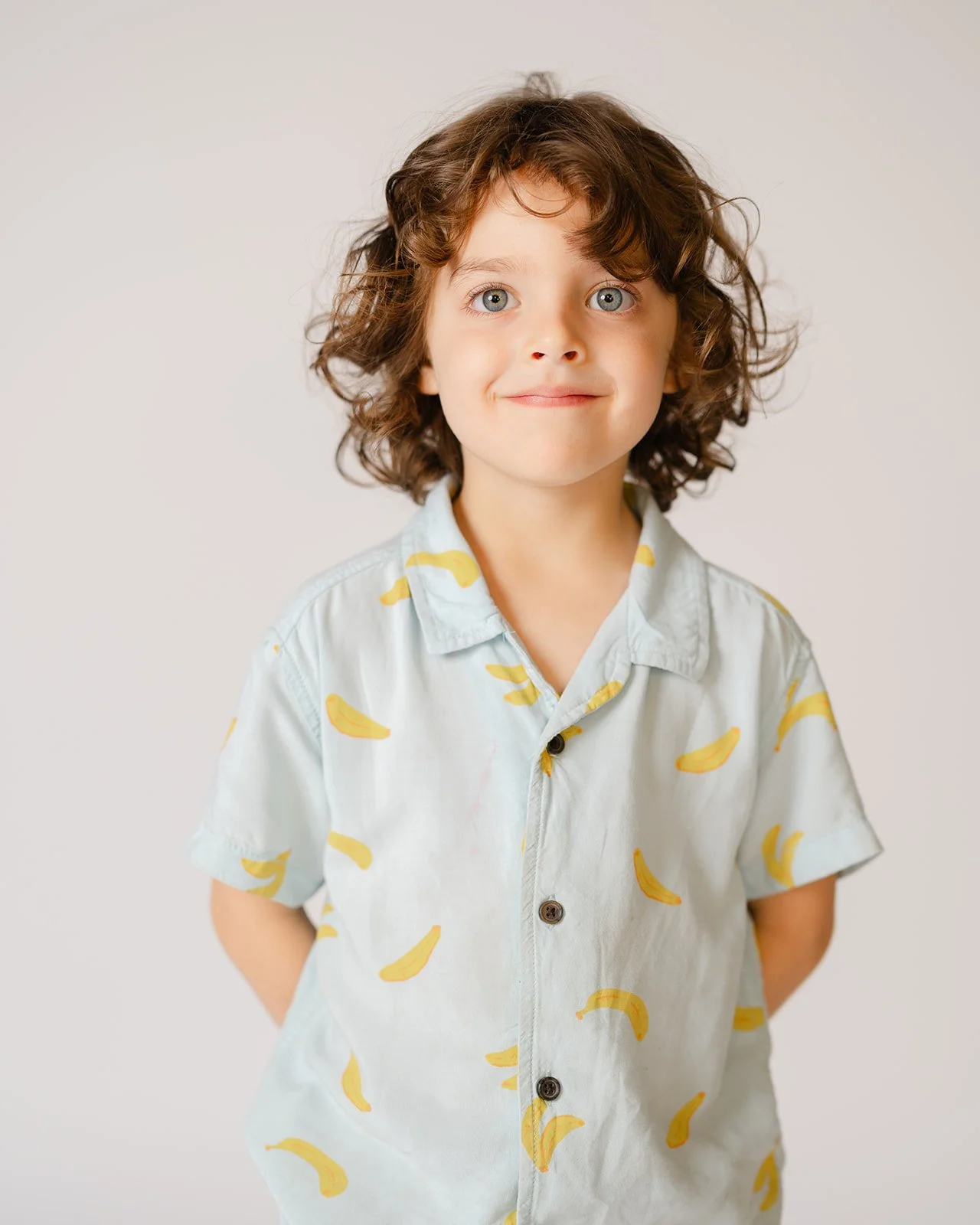A young boy with curly brown hair and blue eyes, wearing a light-colored shirt with a banana pattern, standing against a plain background.