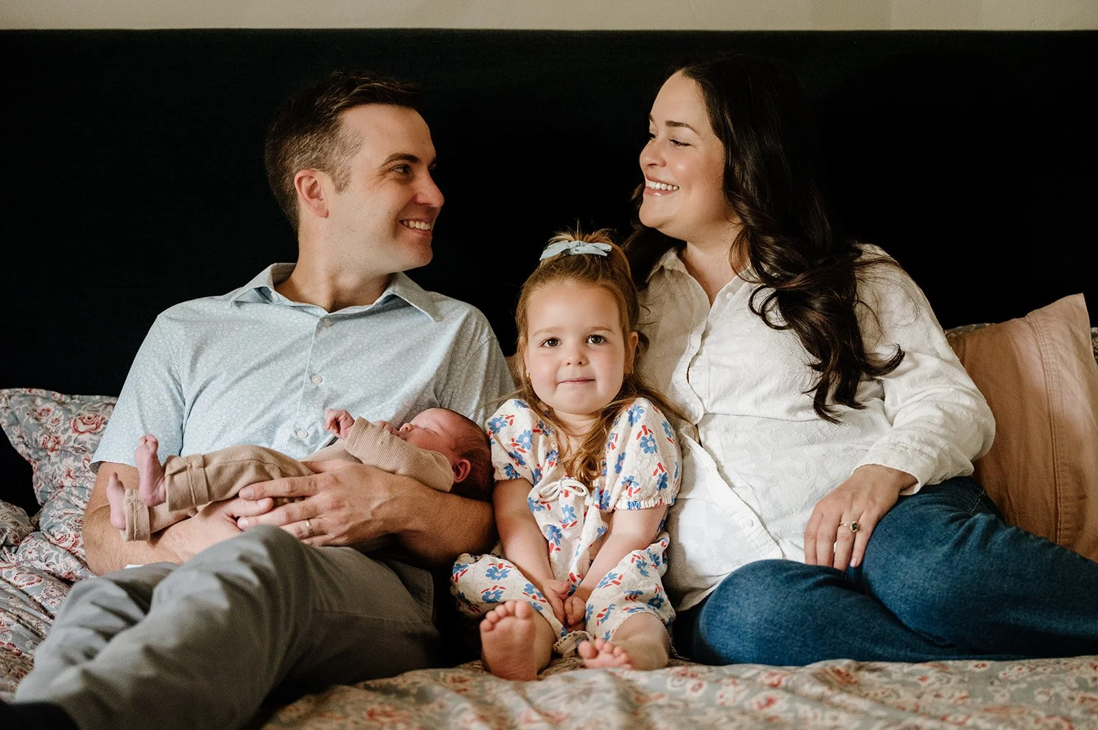 A family of four sitting on a bed, smiling, with a newborn baby. The father holds the baby while the mother and young daughter sit close by, all smiling and looking happy.