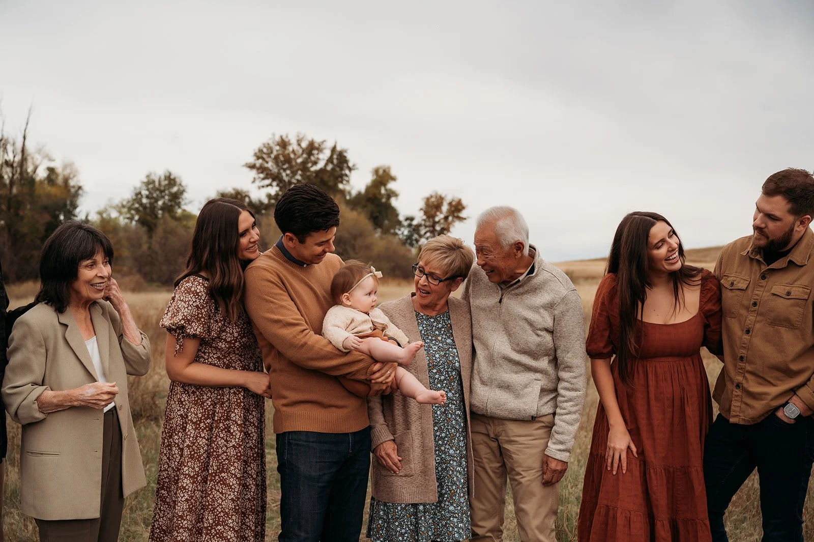 Family gathering outside on a cloudy day, with multiple generations smiling and interacting, including a baby being held by a young man.