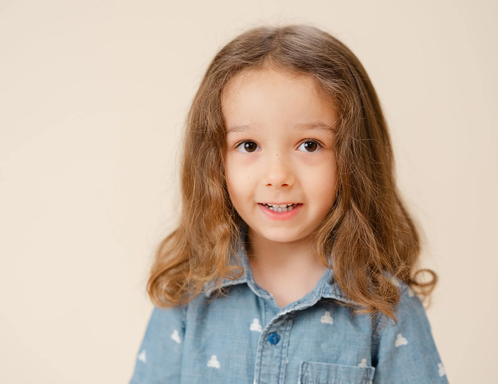 Young girl with long curly brown hair, wearing a denim shirt with white cloud patterns, smiling against a beige background.