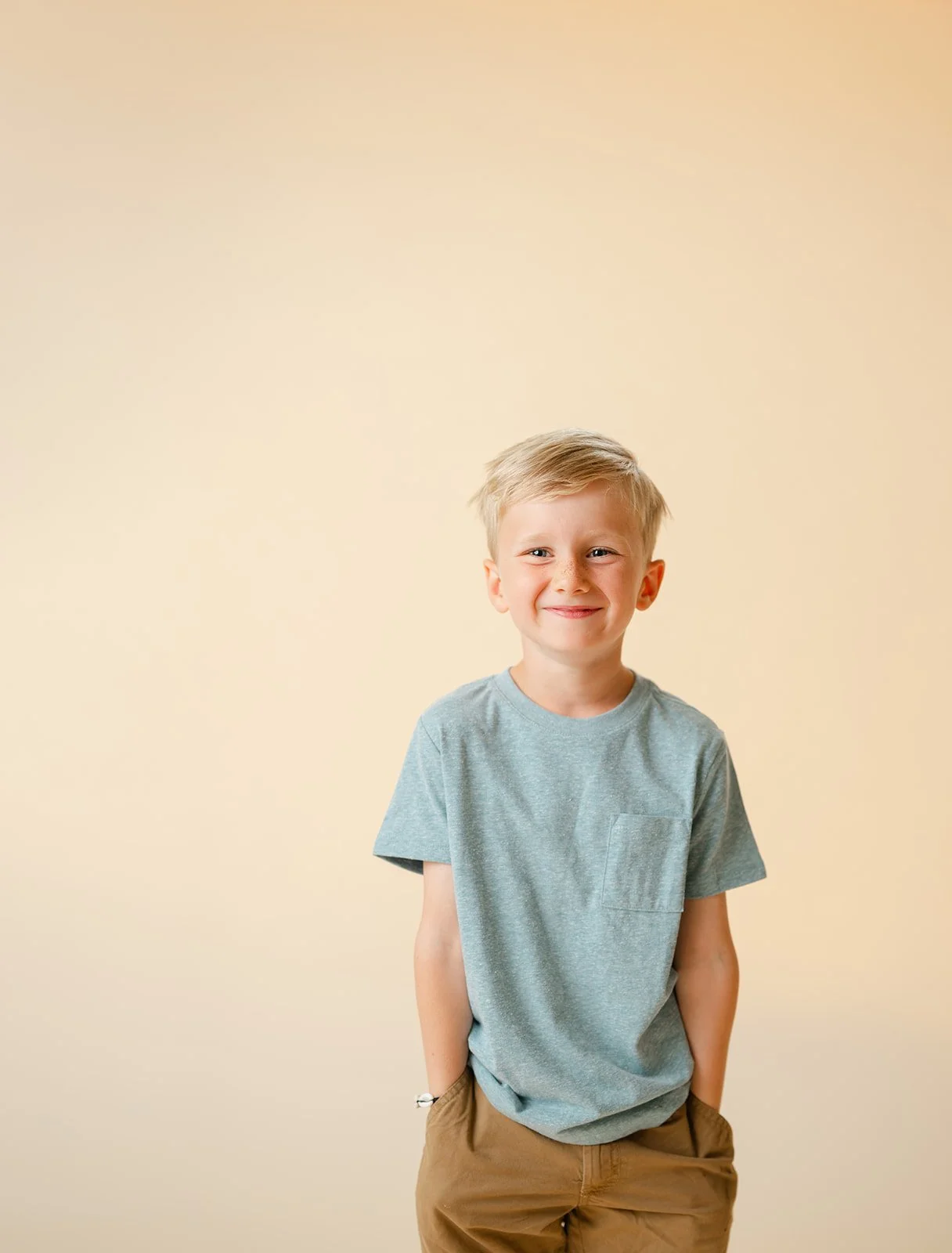 A smiling young boy with blond hair wearing a light blue t-shirt and khaki shorts, standing against a plain beige background.