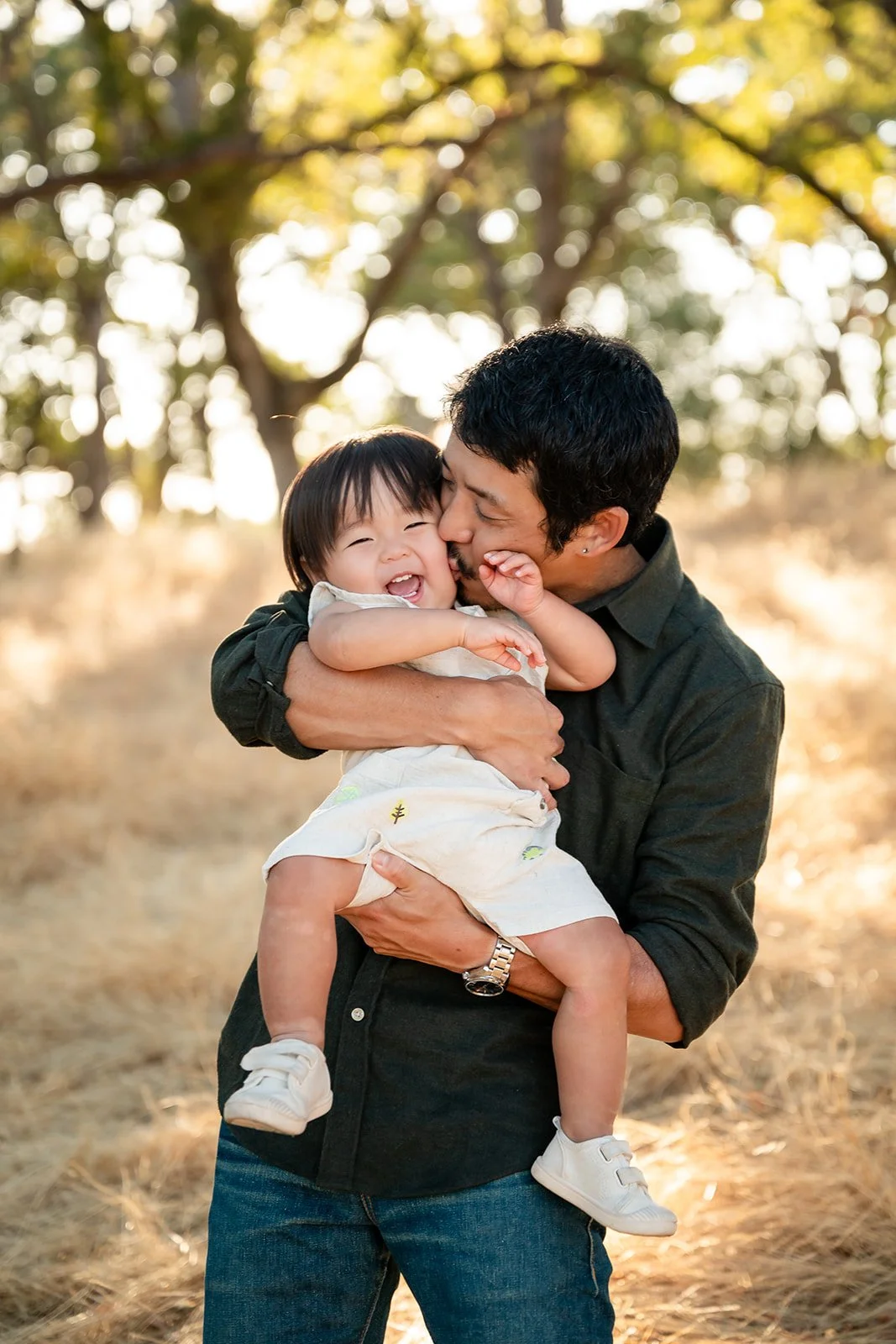 A man is holding a laughing young girl in an outdoor setting with trees and sunlight in the background.