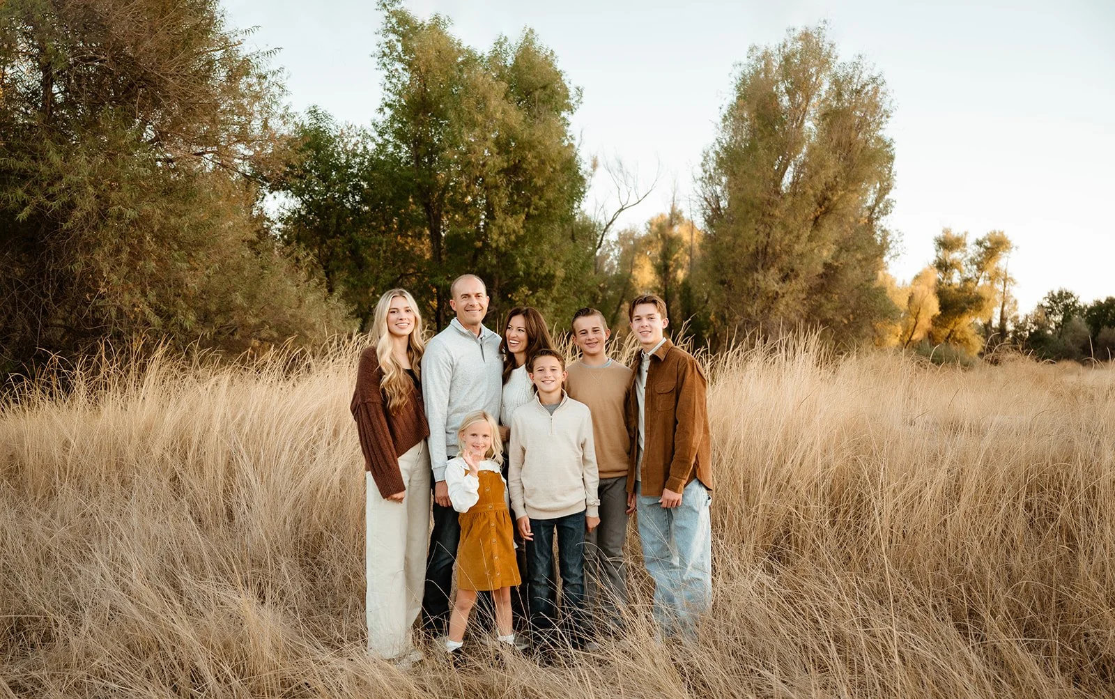 Family of seven members standing in a field of tall, yellowish grass with trees in the background during sunset.