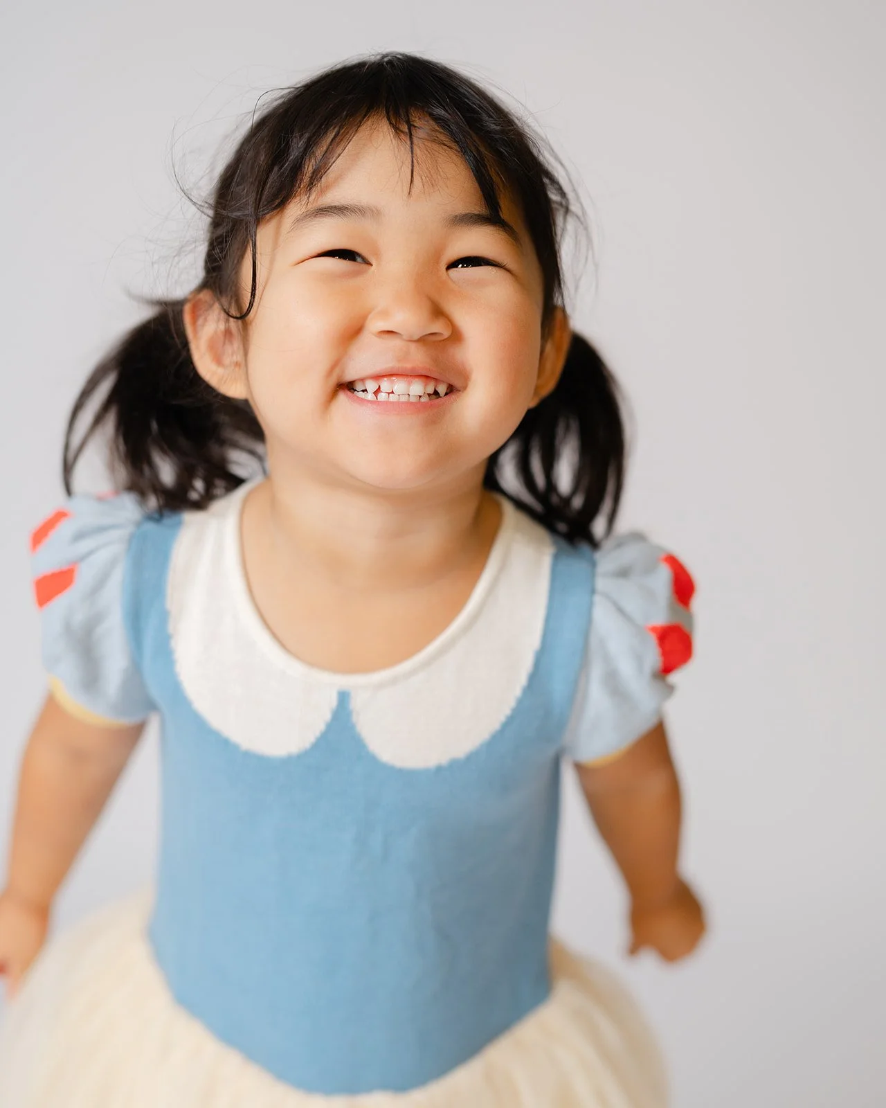 A young girl with black hair styled in pigtails, smiling joyfully at the camera. She is wearing a blue and white dress with scalloped edges and red accents on the sleeves. The background is plain and light-colored.