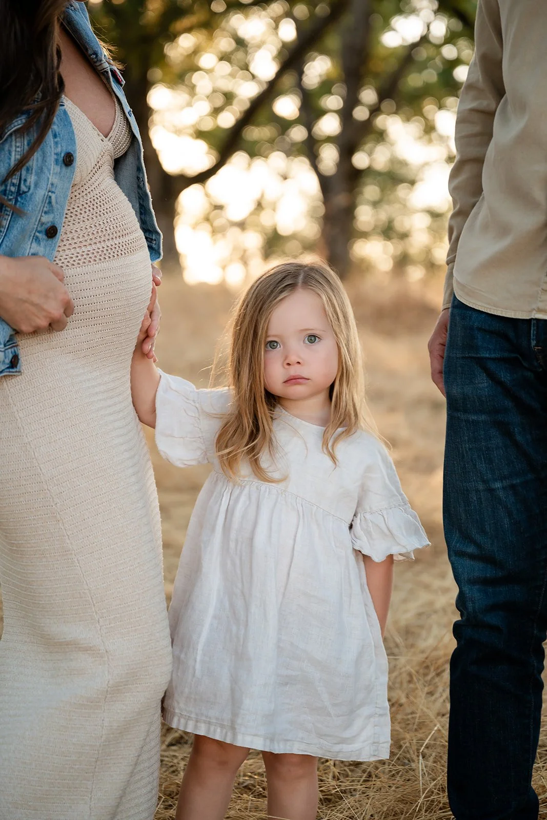 A young girl with long blonde hair and blue eyes, wearing a white dress, stands between her pregnant mother and father outdoors during sunset.