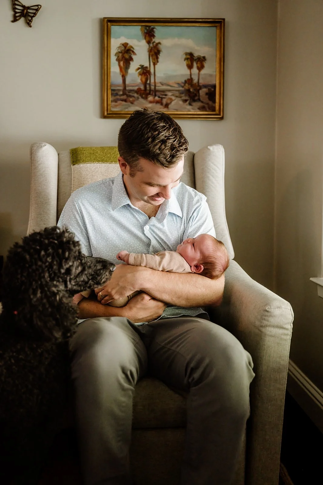 A man sitting in a beige armchair holding a baby, looking down at the baby with a smile. A black curly-haired dog is sitting nearby looking at the baby. A framed painting of palm trees and a coastal scene hangs on the wall behind them.