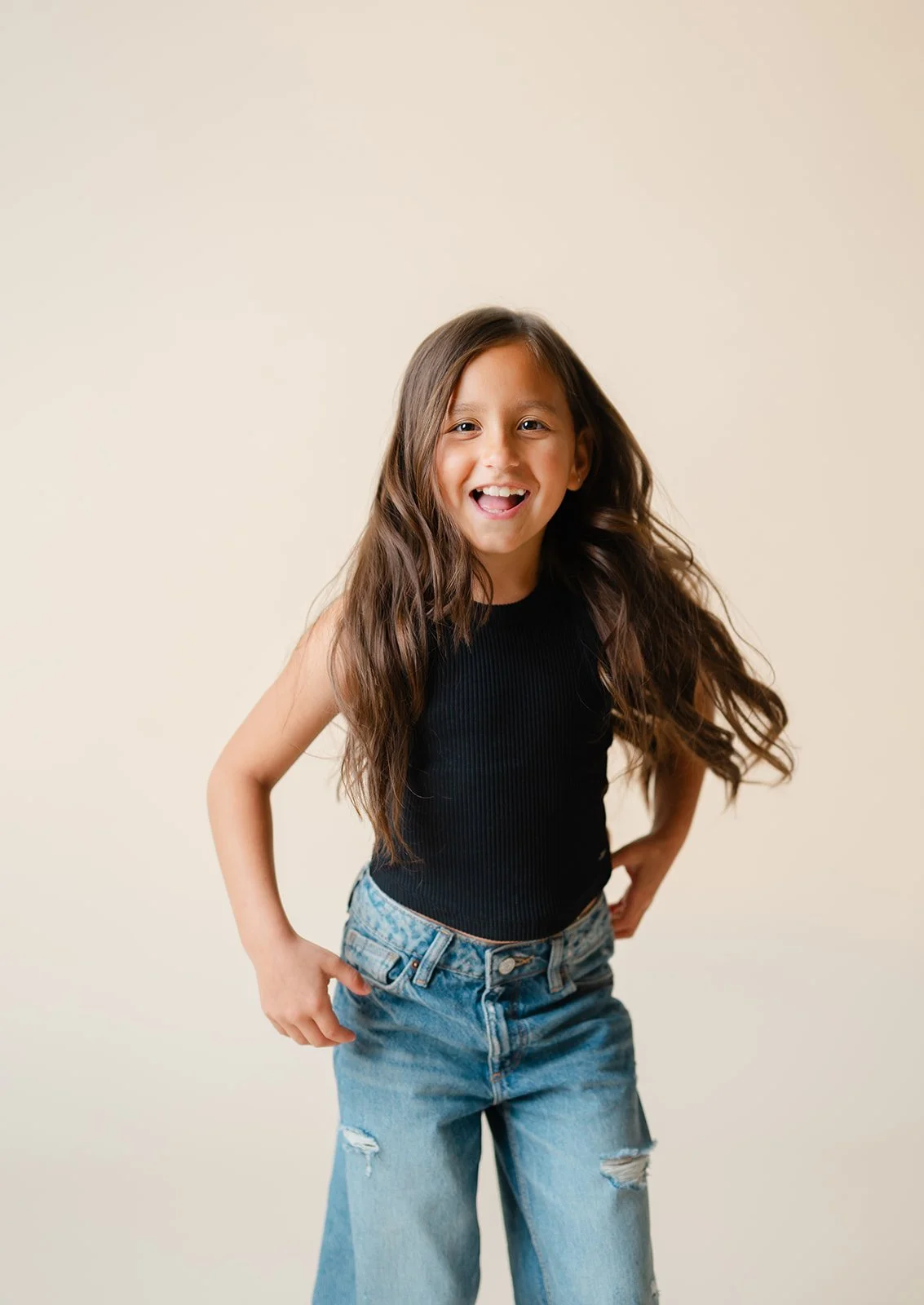 A young girl with long brown hair, smiling, wearing a black sleeveless top and ripped blue jeans, standing against a plain beige background.