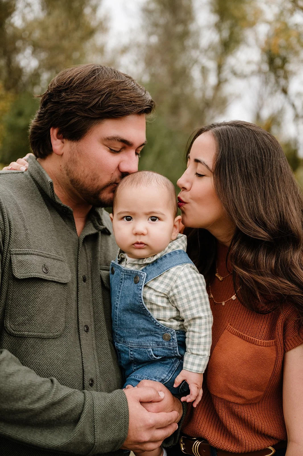 A family of three outdoors, with a man and woman kissing their baby on the cheeks, during fall.