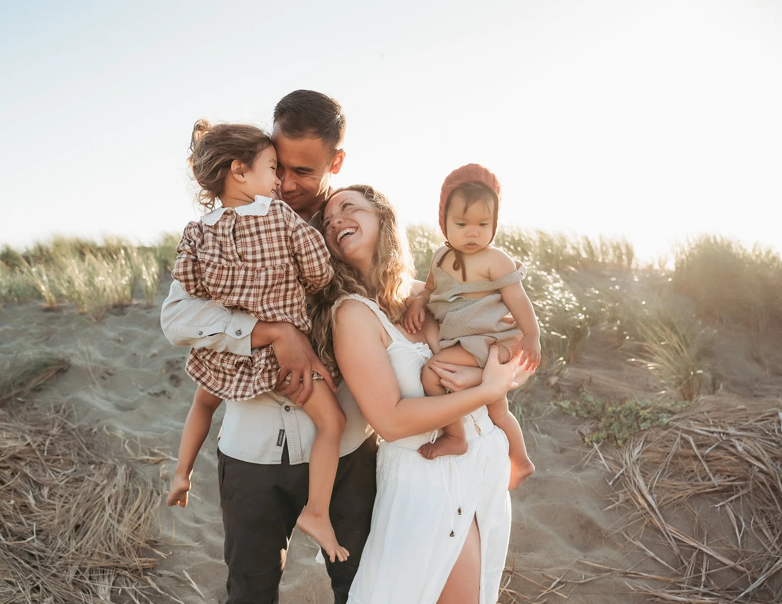Family of four enjoying a moment on a sandy beach during sunset, with two young children, a man and a woman smiling and hugging.