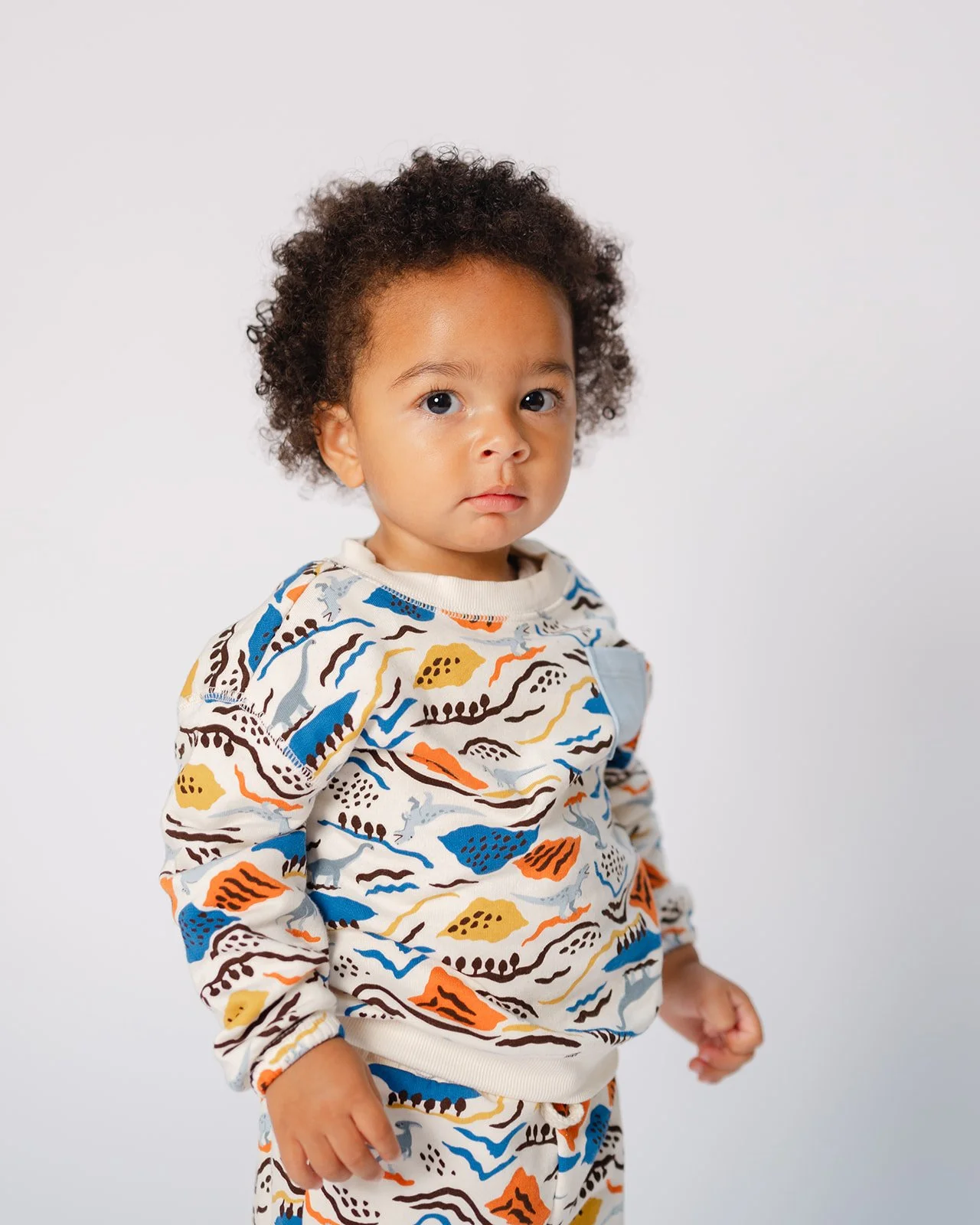 A young child with curly hair and light brown skin wearing a colorful patterned sweatshirt and pants, standing against a plain white background.