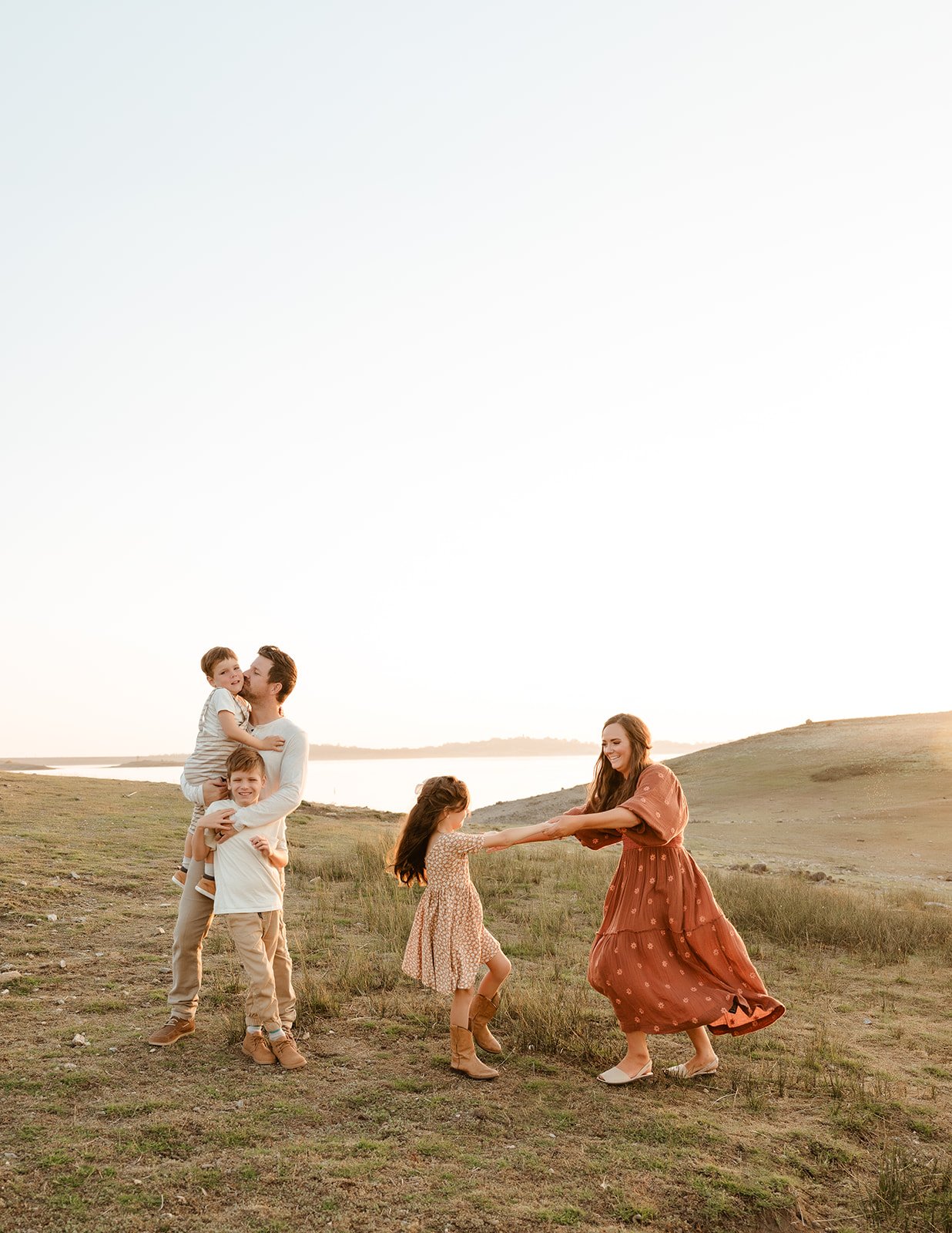 Family of five enjoying a playful moment outdoors at sunset. A woman spins a girl, while a man carries two children on his shoulders near a body of water and green hills.