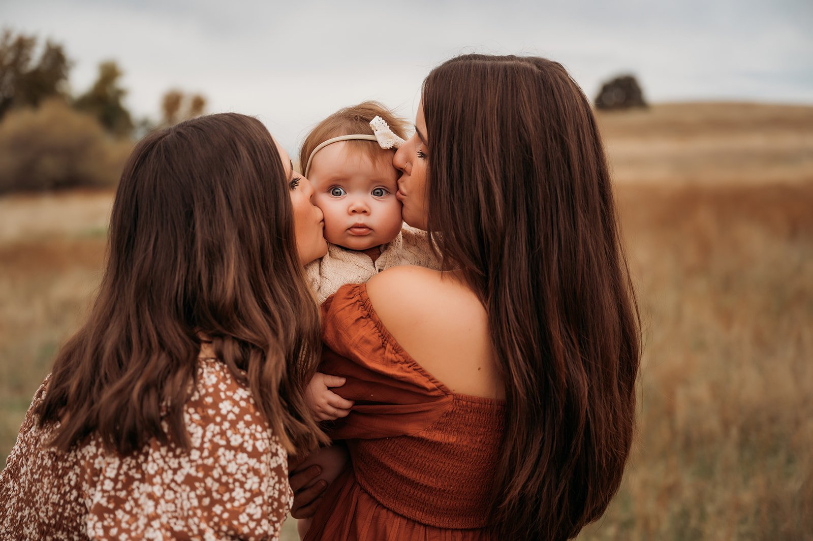 Two women kissing a baby girl on the cheeks in an outdoor field, with a blurred background of trees and sky.