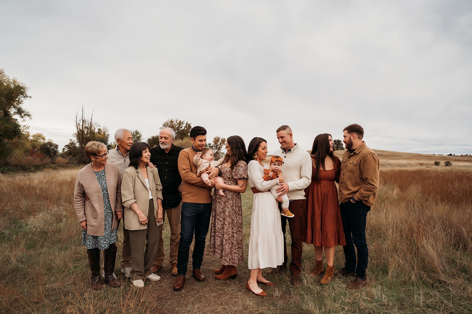 A multigenerational family of fifteen people standing outdoors in a grassy field with trees in the background, smiling and interacting with each other.