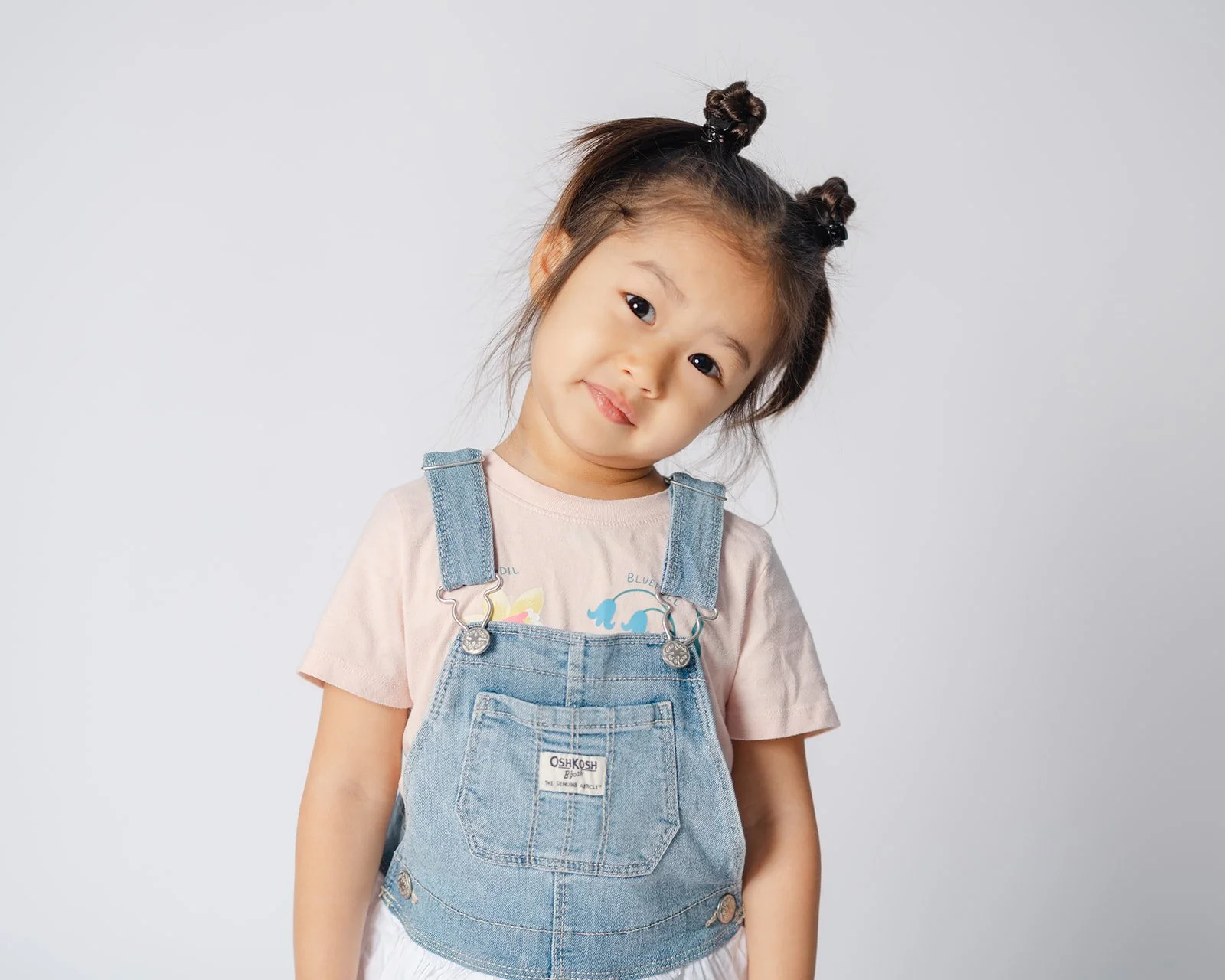 Young girl with dark hair styled in two buns, wearing a pink t-shirt and denim overalls, smiling slightly while tilting her head to one side against a plain white background.