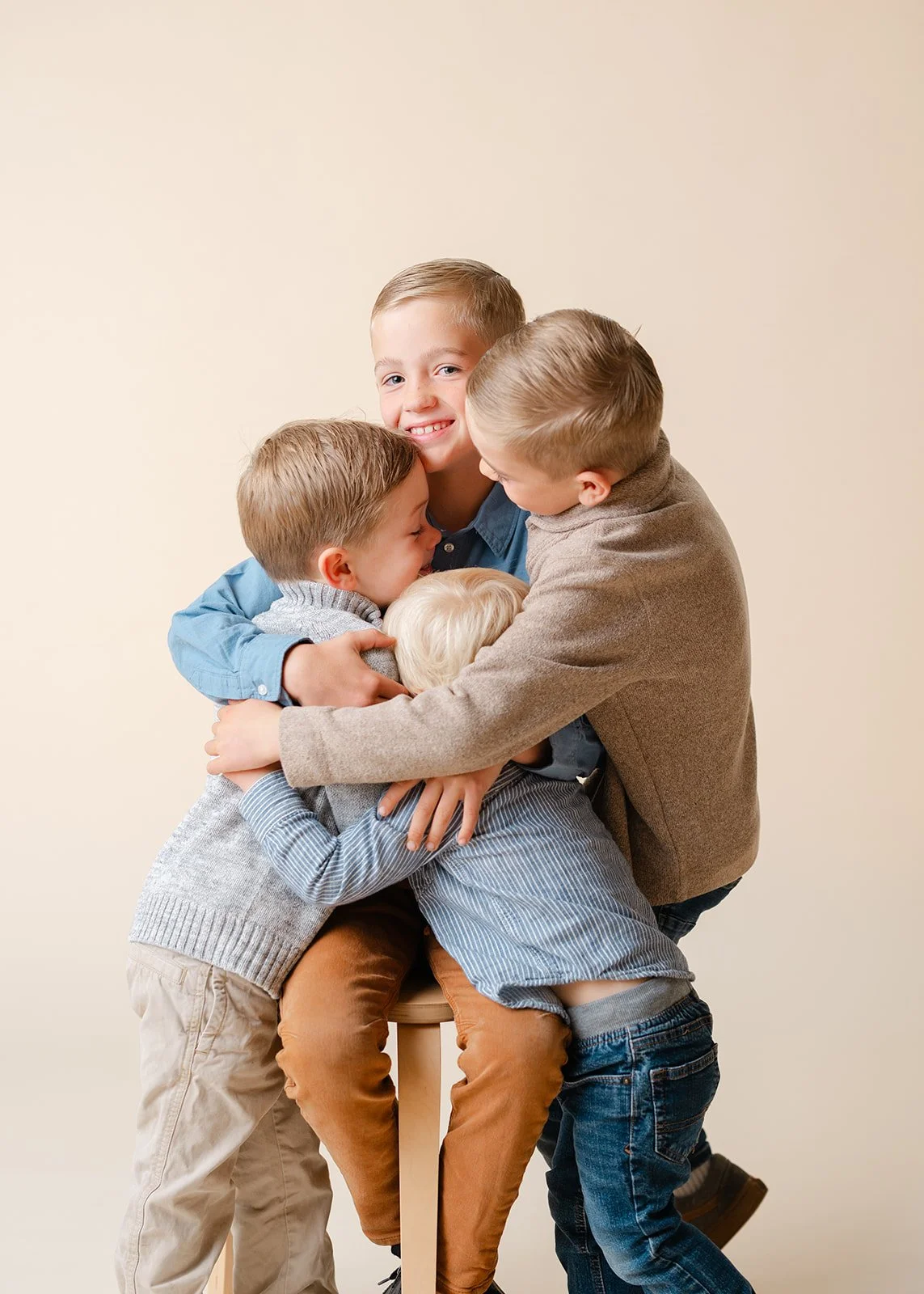 Five children hugging each other in a group hug against a beige background, smiling and showing affection.
